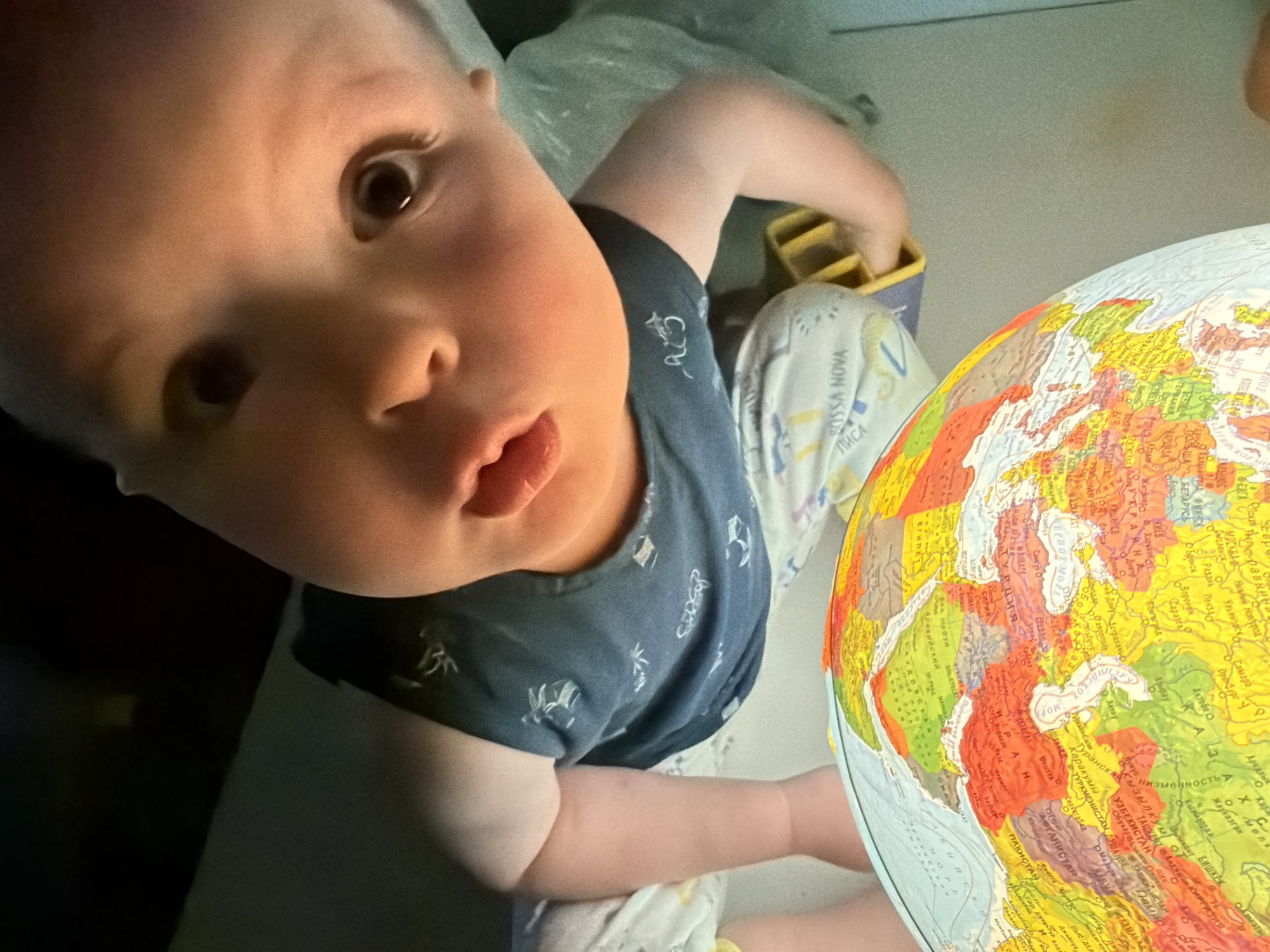 This is a close-up, overhead shot of a baby looking up at the camera, with a globe to their right. The baby is wearing a dark blue t-shirt with a white pattern. Their face is illuminated by a light source coming from the globe. The baby's expression is curious and slightly surprised. The globe shows a map of Europe and Asia, with text in Cyrillic script visible, including names of countries like "ГЕРМАНИЯ" (Germany) and "ПОЛЬША" (Poland), and cities such as "Стамбул" (Istanbul) and "Астрахань" (Astrakhan). The background is dimly lit and appears to be an indoor setting, possibly a room in a home in Votkinsk, Russia. The lighting suggests it could be nighttime or a dimly lit room. The overall mood is intimate and playful, capturing a moment of a child's exploration and curiosity.