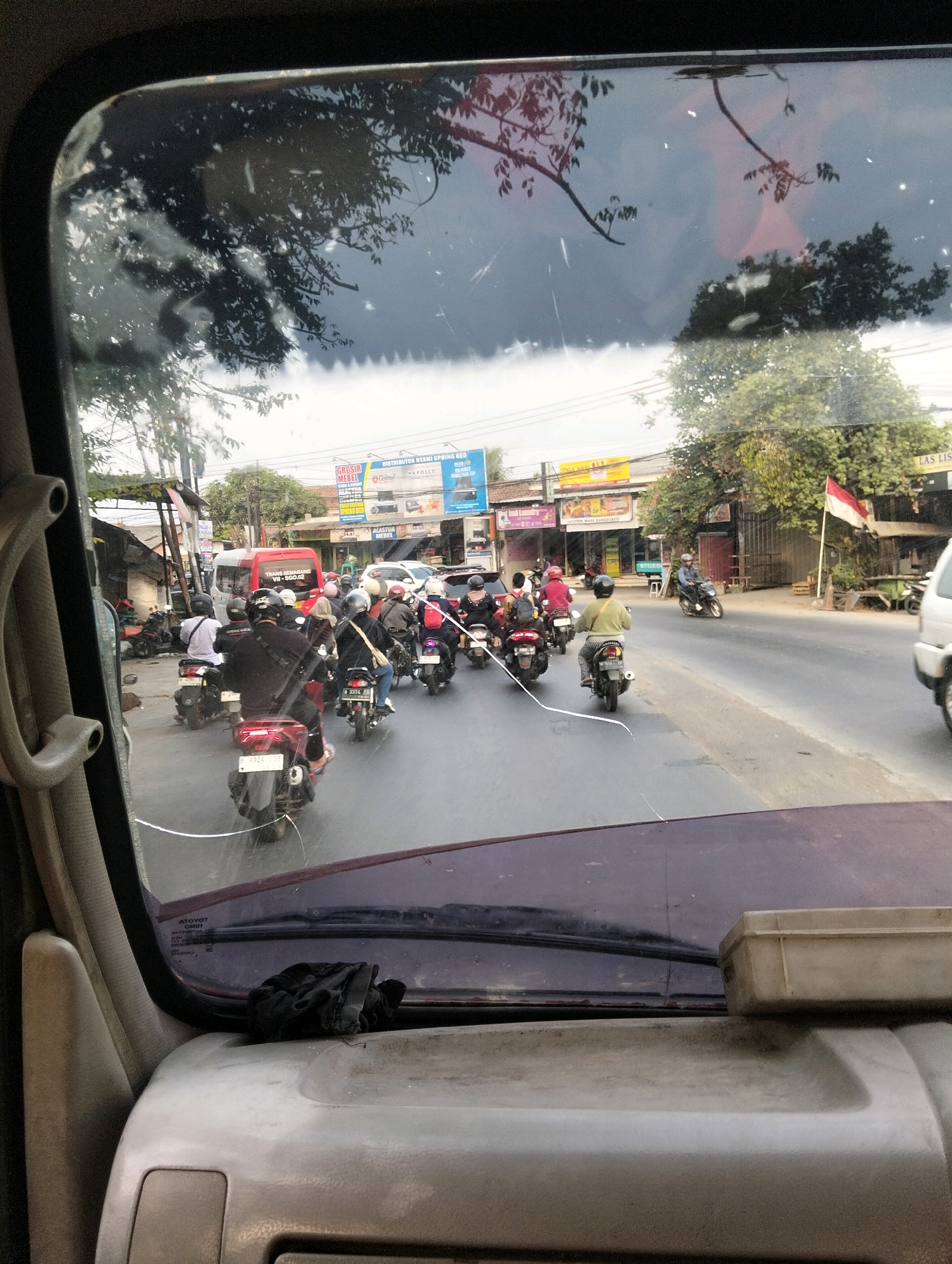 The image depicts a street scene in Semarang, Indonesia, viewed from inside a vehicle. A line of motorcycles, many with riders wearing helmets, is traveling on a paved road. The motorcycles are of various colors, including red, black, and white. Some riders are carrying passengers. In the background, there are buildings lining the street, including shops with signs. One sign appears to advertise "GROSIR MEBEL" (furniture wholesale). A red bus is also visible on the road. A red and white flag is flying on a pole in front of one of the buildings. The sky is overcast, and trees with green foliage are visible. The windshield of the vehicle through which the scene is viewed has several visible cracks and is smudged. A black cloth is resting on the dashboard of the vehicle in the foreground. The overall impression is of a busy urban street with significant motorcycle traffic.