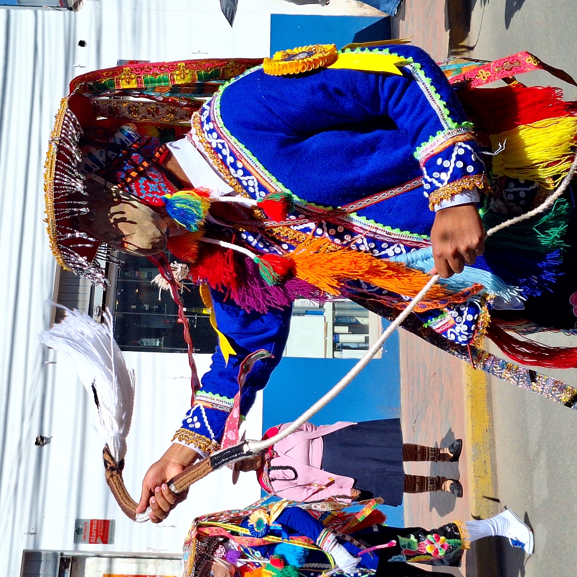 The image shows a person in traditional Wanchaq, Peru attire, likely participating in a festival or cultural event. The subject is adorned in a vibrant blue tunic with ornate, colorful embroidery featuring intricate patterns and gold trim. The costume includes a wide, richly decorated hat with fringes and tassels in yellow, red, and blue. The person is holding a whip-like object with a white feathery end in their right hand, and a white rope in their left hand, suggesting they are performing a dance or ritual.
The background features a white corrugated wall on the left, and a textured paved ground with a yellow curb on the right. Sunlight suggests it is daytime, likely under clear weather conditions. Reflections on the ground indicate a sunny day. The overall scene is lively, implied by the colorful costume and energetic posture. The person's face is partially obscured by the mask and headdress, but the overall impression is one of focused participation.
Below the main subject, another individual in a similarly colorful, albeit less elaborate, costume is visible, suggesting a group performance or procession. There are some shadows cast on the ground, indicating the direction of the sun. A red and white "Alarma" sticker with the Verisure logo is visible on the white wall, indicating a nearby commercial establishment. The context of Wanchaq, Peru, strongly suggests a folk celebration, possibly a religious or agricultural festival.