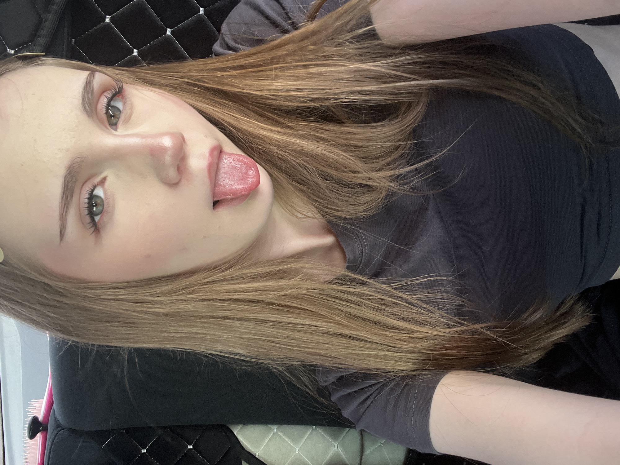 A young woman with long, light brown hair is pictured from the chest up, taking a selfie. She is wearing a dark grey t-shirt. Her right eye is looking directly at the camera, while her left eye is looking slightly off to the side. Her tongue is sticking out. She has light makeup on, including pink blush and mascara. The background appears to be the interior of a vehicle, with a black quilted seat visible to the right of the woman and a lighter patterned seat to the left. There is a pink hairbrush with a black handle on the left side of the frame, resting on the black seat. The location is identified as Novaya Otradovka, Russia.