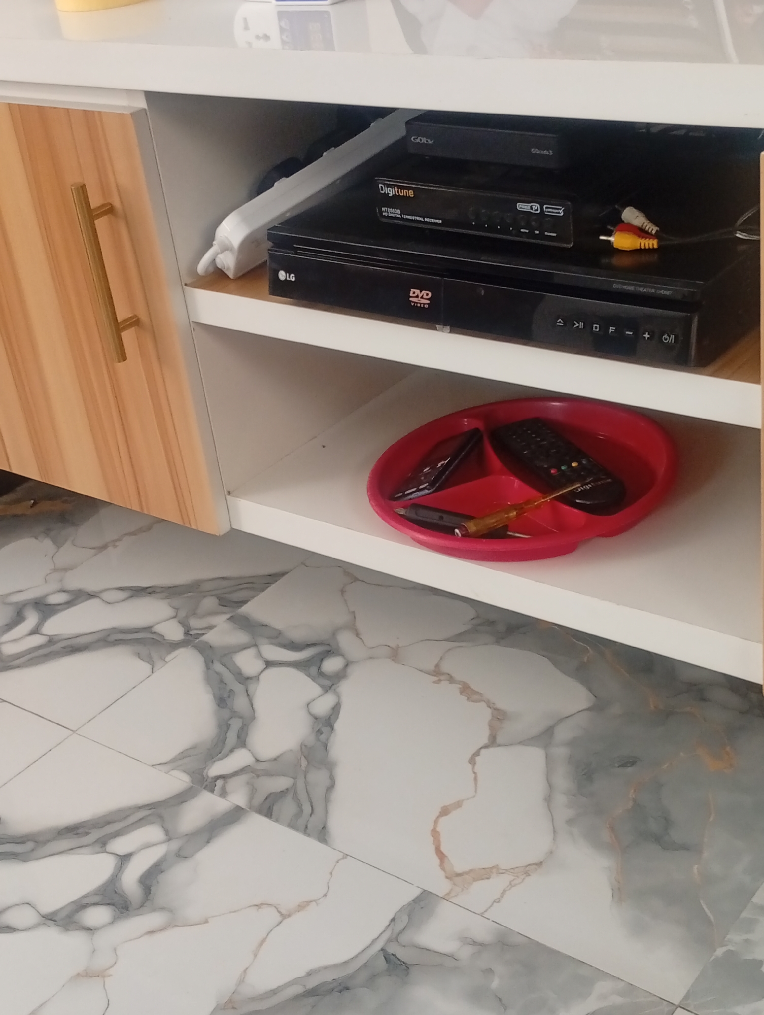 A medium shot captures a close-up of a wooden cabinet with a brass handle on the left side of the frame. The cabinet is open, revealing two shelves. The top shelf contains a black "GOtv" digital receiver. Below it, on the second shelf, are a black "Digitune" HD digital terrestrial receiver and a black "LG" DVD player. RCA cables are connected to the Digitune receiver. In the center of the second shelf, a red plastic tray holds two remote controls, a black mobile phone, and a yellow-tipped screwdriver. The cabinet is positioned above a marble-patterned tiled floor. The scene appears to be indoors, likely in a home environment. The exact location of Rukuba Road, Nigeria, is not visually discernible from the image itself.