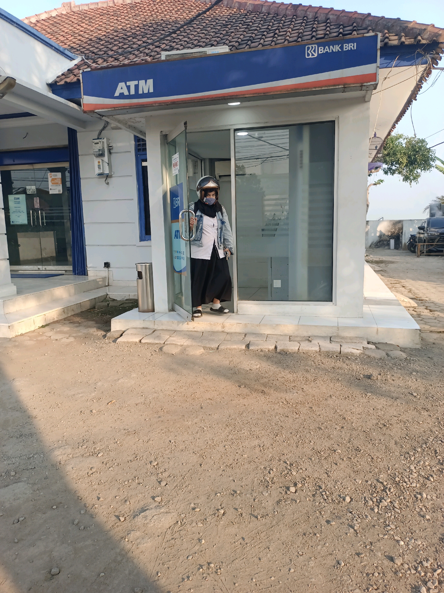 The image depicts an ATM booth for BANK BRI located in Subang, Indonesia. A person wearing a hijab, a denim jacket, and black pants is standing inside the ATM booth. The person is also wearing a helmet and a face mask. The ATM booth has a glass door and a blue and orange sign that says "ATM" and "BANK BRI". There is a trash can next to the booth. The ground in front of the booth is covered with dirt and gravel. In the background, there is a building with a tiled roof and some trees. A car and a motorcycle are parked nearby. The sky is clear and blue, suggesting it is daytime.