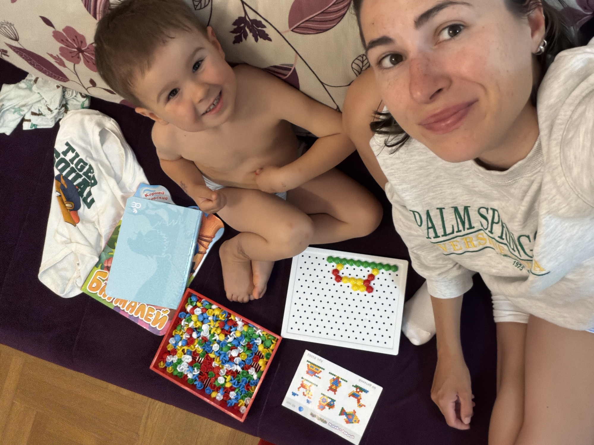 A medium shot captures a woman and a young child in an indoor setting. The woman, positioned on the right, has fair skin, brown hair, and is wearing a light gray sweatshirt with the words "PALM SPRINGS" in green. The child, situated to the left of the woman, has fair skin, short brown hair, and is wearing a diaper. They are both smiling and looking towards the camera.
Scattered around them on a dark purple surface are various objects, including a white t-shirt with "TIGER SQUAD" printed on it, a children's book with Cyrillic text, a light blue notebook, a red box filled with colorful plastic shapes, a white perforated board with some plastic shapes inserted, and an instruction sheet with diagrams of shapes.
The background includes a patterned pillow with purple and white floral designs. The setting appears to be a casual, domestic environment, possibly a living room or play area. It is not possible to identify the location as the city of Budva, Montenegro from the visual information provided.