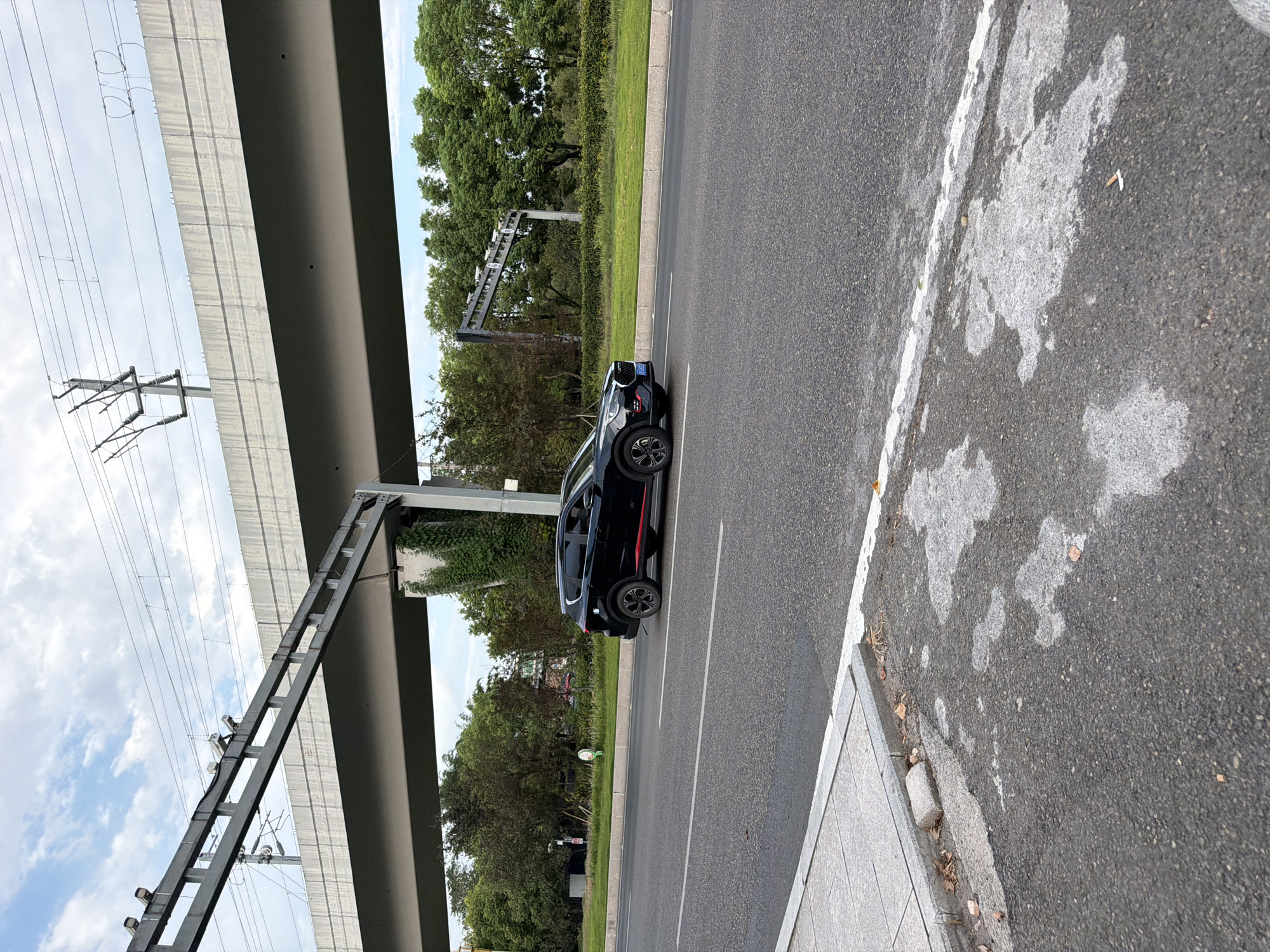 In Jiangjin, China, on a paved road under an elevated highway structure, a black SUV with red trim is captured in motion. The SUV is traveling on the right side of the road, with its front facing towards the left of the frame. The road surface appears wet in patches, and a white line marks the lane. To the left of the road, a grey sidewalk with a concrete curb is visible. Beyond the road, a grassy median separates it from trees and vegetation. The elevated highway features a grey concrete structure with overhead gantries supporting traffic cameras. Power lines and poles are present above the highway. The sky is partly cloudy.