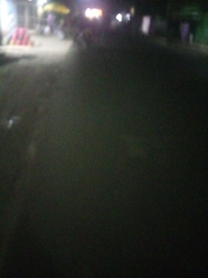 The image captures a poorly lit outdoor scene at night. The foreground is dominated by a dark, textured surface, possibly a road. In the distance, blurred lights and indistinct shapes suggest a building or structure, with hints of activity or parked vehicles. The overall mood is one of darkness and obscurity, making it difficult to discern specific details.