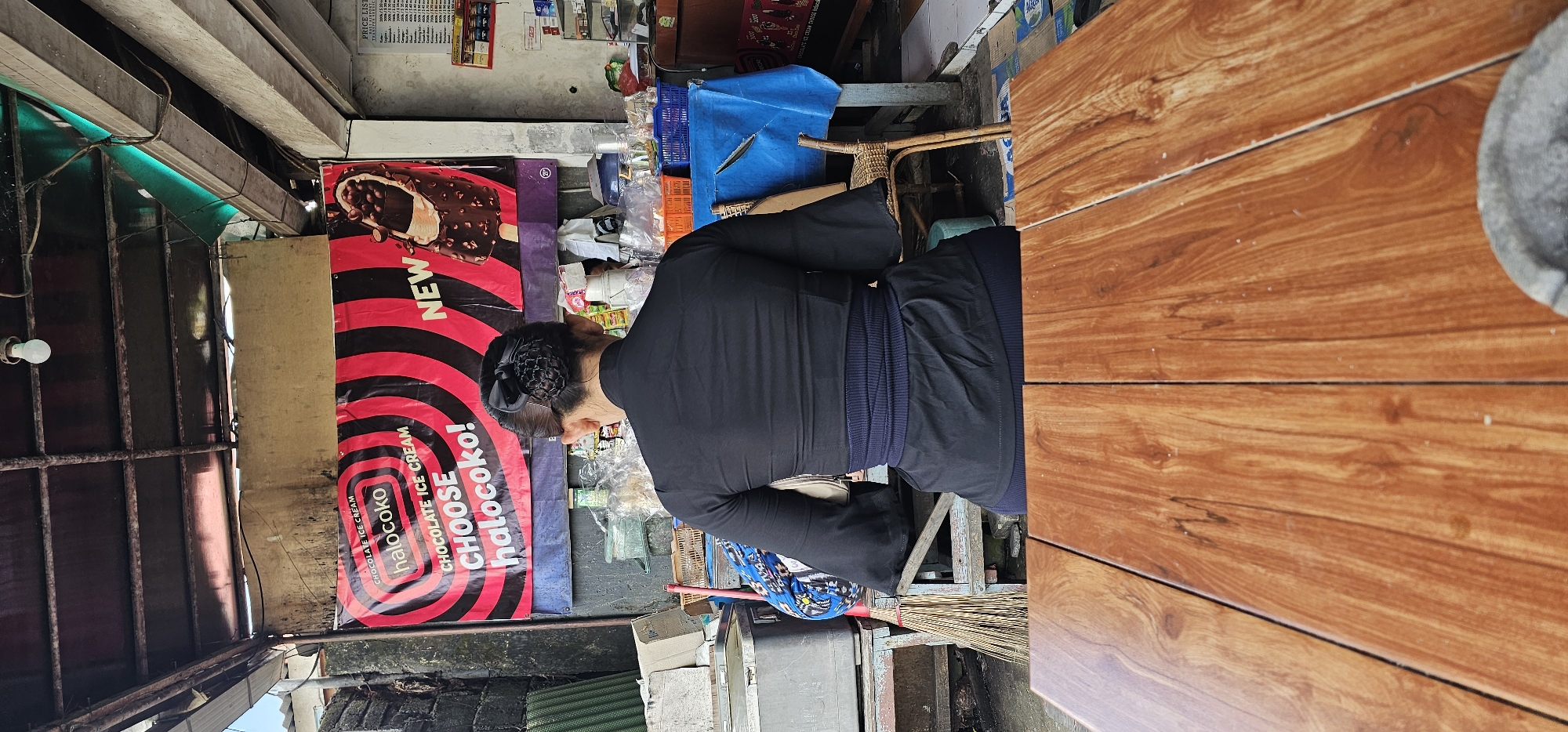 A person, seen from the back and right side, stands or sits at what appears to be a stall or small shop. They are wearing a long-sleeved black top and dark trousers, with their dark hair neatly tied back in a bun secured by a net. To the person's left, a large red and black banner advertises "halocoko" chocolate ice cream, featuring an image of an ice cream bar and the words "NEW," "CHOCOLATE ICE CREAM," and "CHOOSE halocoko!".
On the right side of the image, a wooden table with a distinct wood grain pattern is prominently featured, with a grey mortar partially visible on its far edge. Behind the person, various shop items are cluttered, including blue plastic crates, items wrapped in clear plastic, and a blue tarp. In the background, a price list is attached to a wall, though its details are unreadable.
The setting appears to be an open-fronted or semi-outdoor stall, characterized by metal grates and exposed wiring on the left side, and a bare light bulb hanging down. A small broom made of natural fibers rests near the ground. The scene suggests a bustling, everyday commercial environment, typical of street vendors or small local shops. The time of day appears to be daylight, providing natural illumination to the scene.