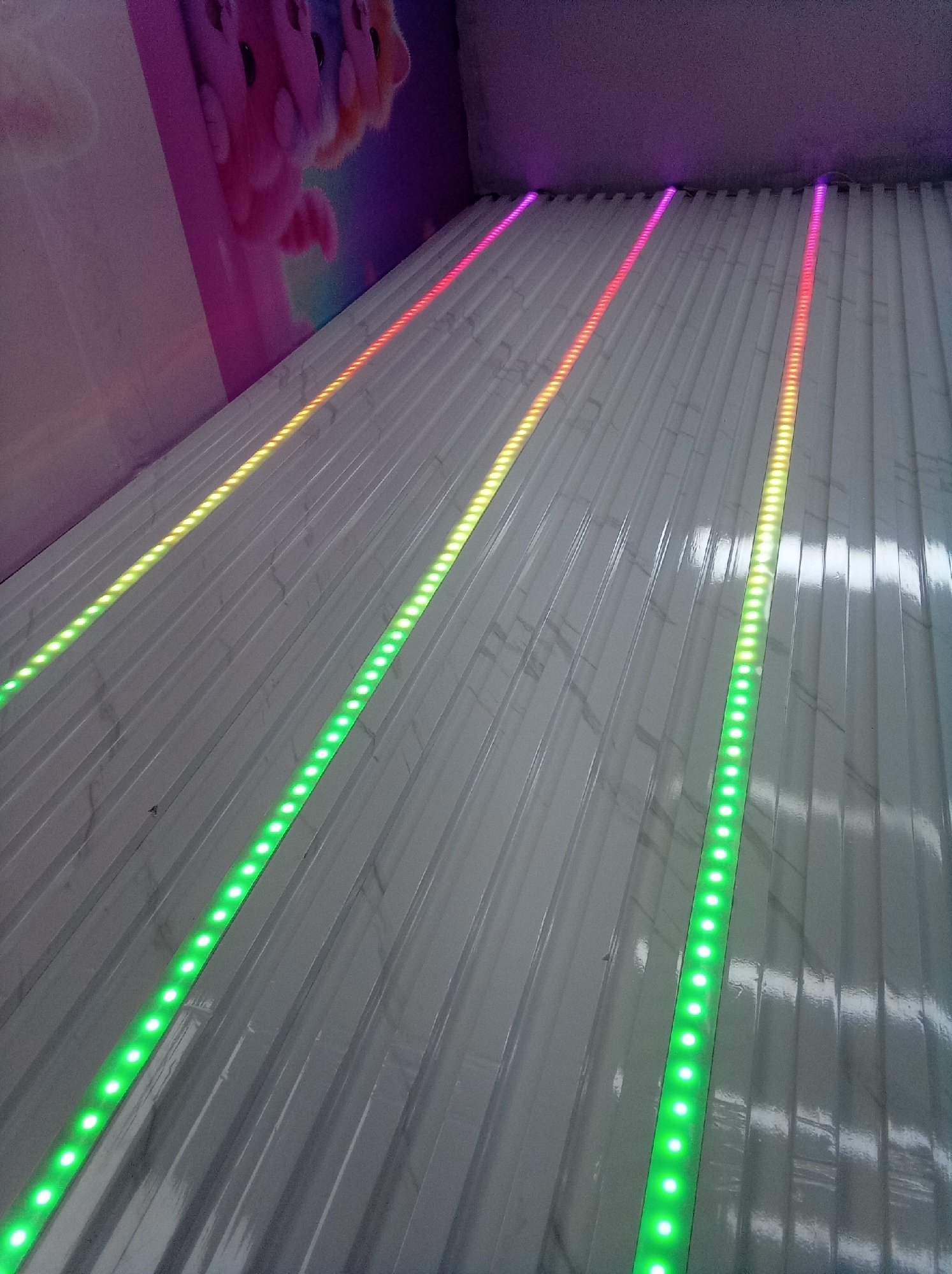 The image shows a view of an indoor slide or ramp with three illuminated LED strips running along its length. The LED strips display a color gradient, changing from green at the bottom to yellow, red, and purple towards the top. The surface of the slide appears to be made of a light-colored, possibly white or gray, material with subtle streaked patterns. A wall is visible on the left side of the frame with a colorful image on it. The lighting is dim overall, with the LED strips providing the primary source of illumination. The location is inside an amusement park in the city of Guarenas, Venezuela.
