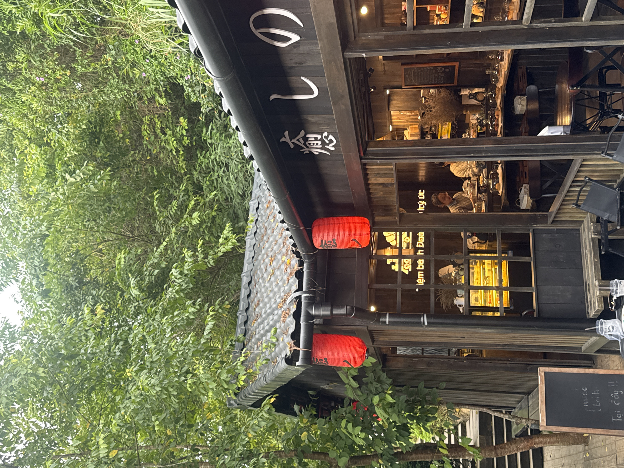 The image shows a Japanese-style cafe in Cam Ly, Da Lat, Vietnam. The cafe has a dark wooden exterior with a traditional tiled roof. Red lanterns hang from the eaves, and Japanese characters are visible on the building's facade. Through the windows, one can see display cases filled with pastries and cakes, as well as customers and staff. The cafe is nestled amongst lush greenery, suggesting a natural or park-like setting. A chalkboard sign is visible outside, and outdoor seating is also present. The overall atmosphere appears to be quaint and inviting.
