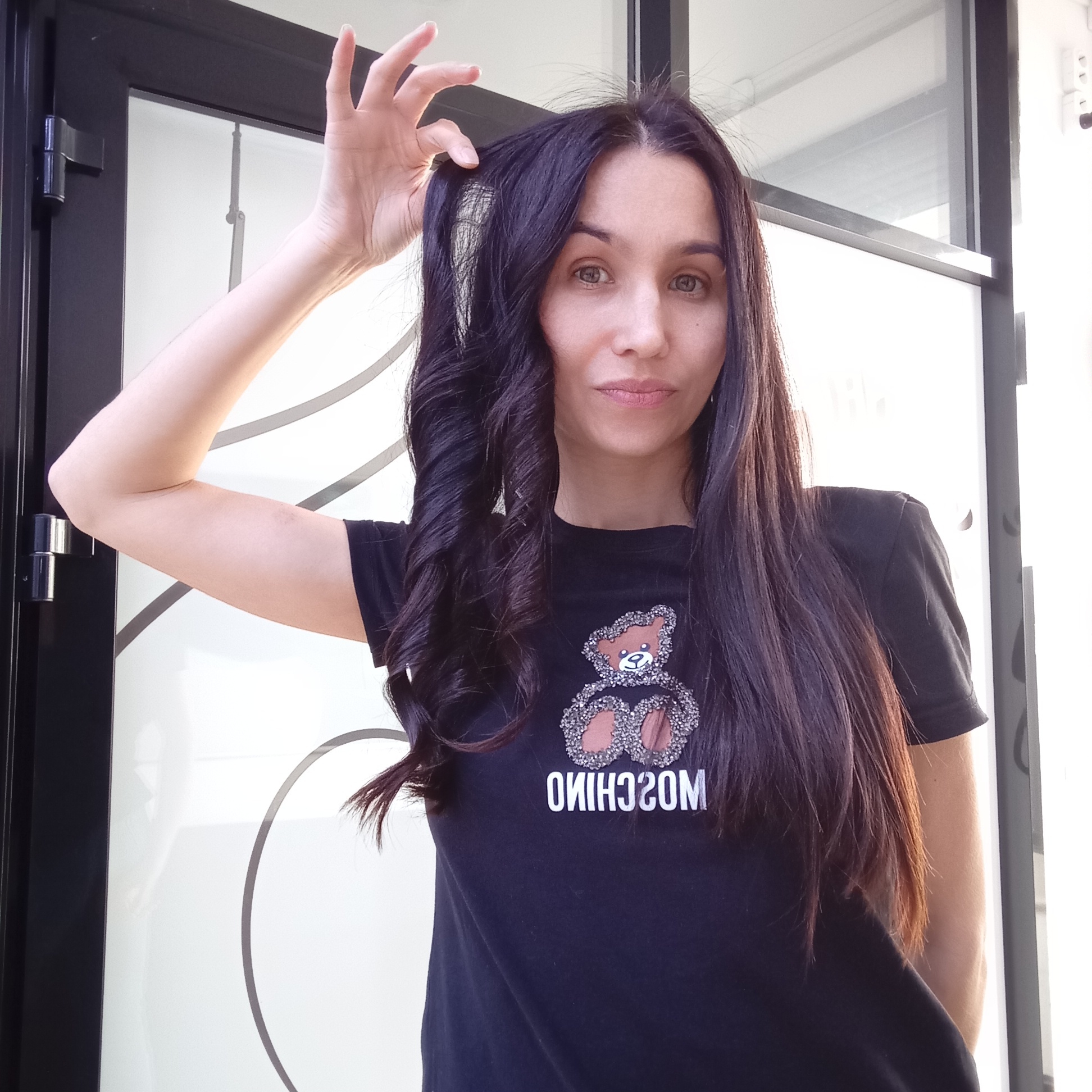 A woman is standing in front of a building in Izhevsk, Russia. She is wearing a black T-shirt with a teddy bear design and the word "MOSCHINO" written backwards underneath it. The woman has long, dark hair, styled with some curls on one side. She is holding a strand of her hair up with her hand. The building behind her has a white door with a black frame, featuring a circular design. The overall lighting suggests it is daytime.