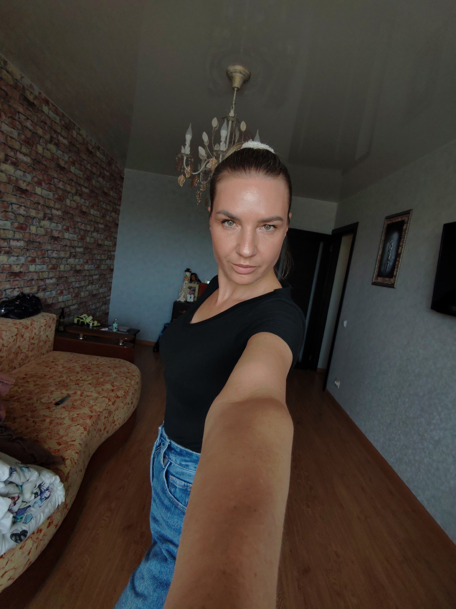 An adult female is captured in a selfie photograph, taken indoors in a residential setting in Petropavlovsk-Kamchatsky, Russia. The subject, appearing to be in her 30s or 40s, holds the camera with her right arm extended towards the viewer. She has light-colored eyes and her hair is pulled back, secured by a white, textured hair tie. She is wearing a black V-neck t-shirt and blue denim jeans. Her expression is neutral, and she looks directly into the camera.
The room features a diverse wall treatment: the left wall is covered in reddish-brown faux brick cladding, while the upper portion of the back wall beyond the sofa is painted a light blue or grey. The right wall is covered in a light grey, textured wallpaper. The ceiling is a uniform light grey or off-white, possibly a stretch ceiling, showing slight reflections and a visible seam. The flooring throughout is a warm brown laminate or parquet.
Furniture includes a large, U-shaped or curved sofa upholstered in a patterned fabric of beige, red, and orange tones, positioned on the left side of the room. A small, rectangular wooden coffee table with a lighter top stands in front of the sofa, holding a dark bottle, a light-colored bottle, and other small items. A dark object, possibly a helmet, rests on the sofa near the left edge, and a crumpled white blanket with a light pattern is visible further along the sofa. A dark, pen-like object lies on a sofa cushion in the foreground.
A multi-armed, cream-colored chandelier with golden leaf-like embellishments and candle-shaped light fixtures hangs from the center of the ceiling. On the right wall, a dark-framed artwork depicting a light central figure is mounted above a white electrical outlet or switch. To the right of the artwork, a dark flat-screen television is wall-mounted. Two dark-colored interior doors are visible in the background on the right, one slightly ajar. The room appears well-lit, likely by natural light from an unseen source, with shadows present under the chandelier and in room corners.