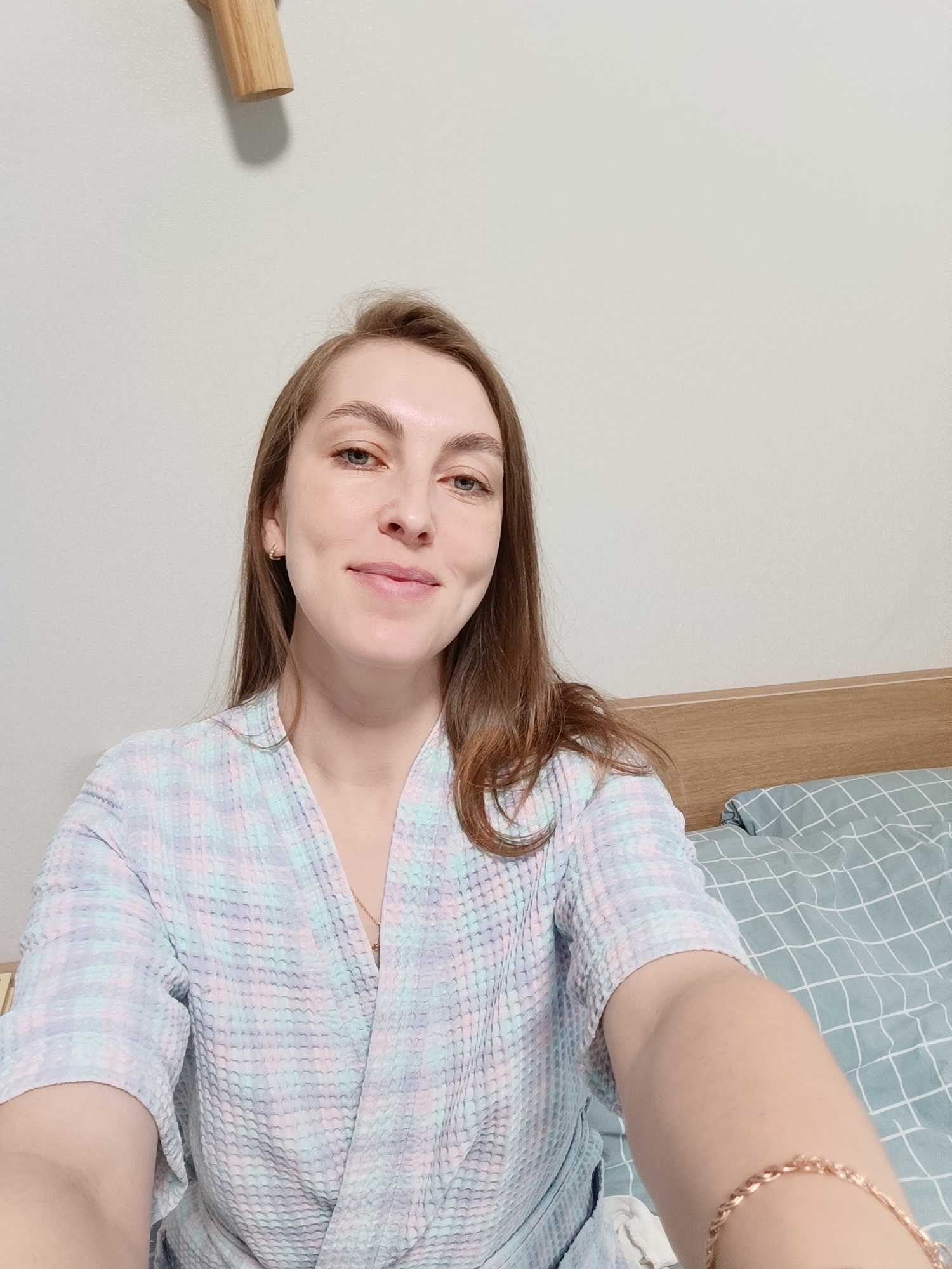 A woman is in a room in Chaikovsky, Russia. The room includes a bed with a patterned blue and white cover, a wooden headboard, and a light-colored wall. The woman has light brown hair and is wearing a light-colored plaid robe. She is smiling slightly. There is a wooden peg on the wall behind her. The image appears to be a selfie. A gold bracelet is visible on her wrist.