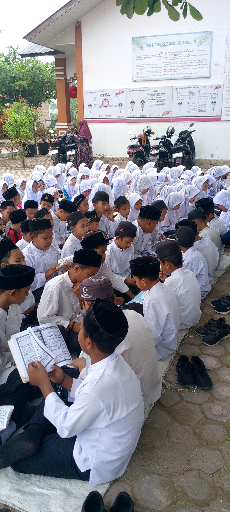 In the city of Baratpayaitek, Indonesia, a group of young students, dressed in white Islamic attire with black caps, are gathered outdoors on a paved surface, possibly for a lesson or religious activity. They are seated on a mat, with some holding open books. The background shows a white building with signage, suggesting it might be a school. Several motorcycles are parked nearby. The overall scene conveys a sense of community and learning.