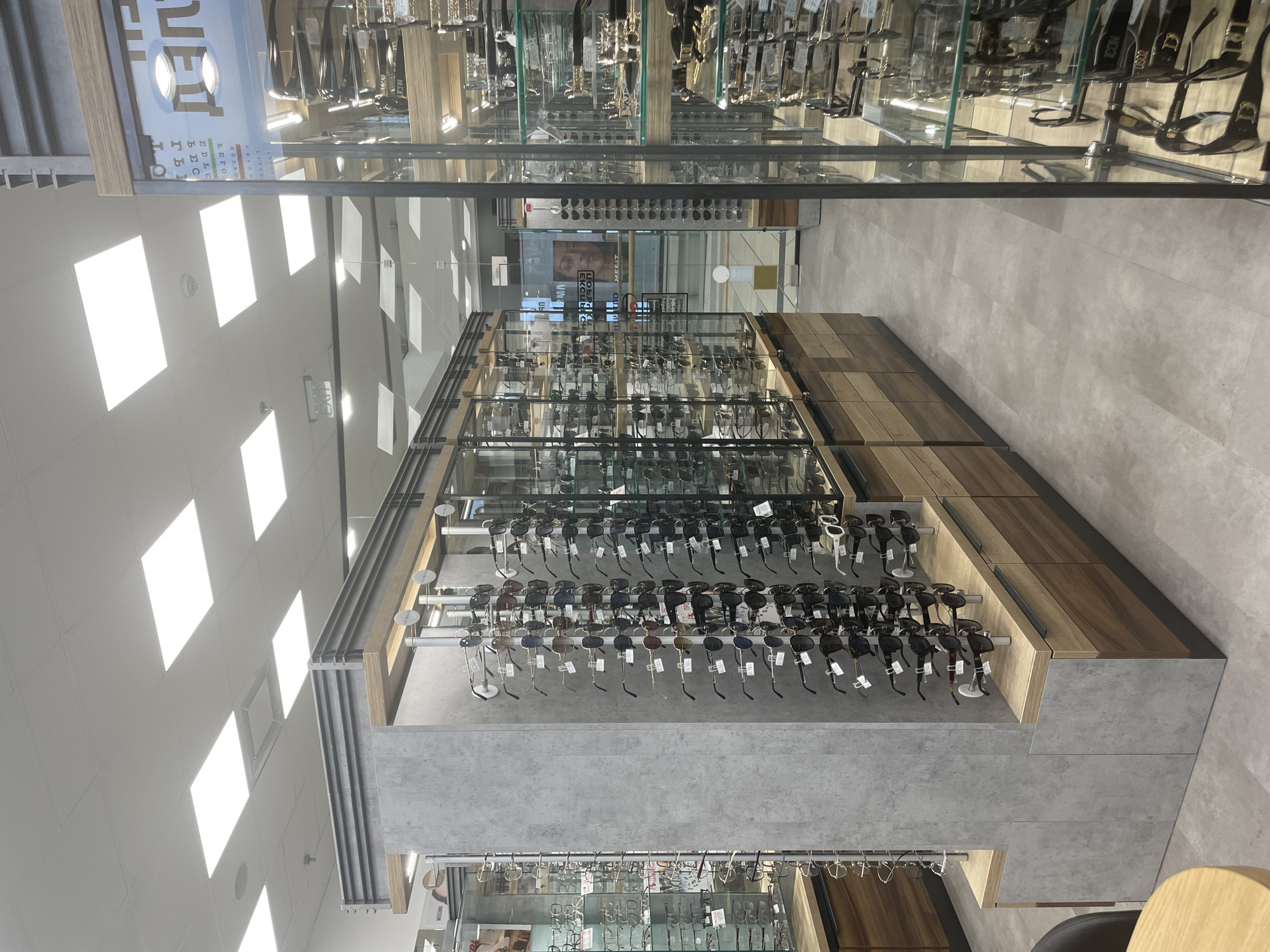 The image depicts the interior of an eyeglasses store, specifically showcasing display shelves filled with eyeglasses and sunglasses. The shelves are constructed from a combination of materials including wood, metal, and gray concrete-like panels. The eyeglasses are arranged on metal rods extending from the shelves. The store has a brightly lit interior with recessed lighting. The floor is a neutral gray tile. The image was most likely taken in Sofia, Bulgaria.