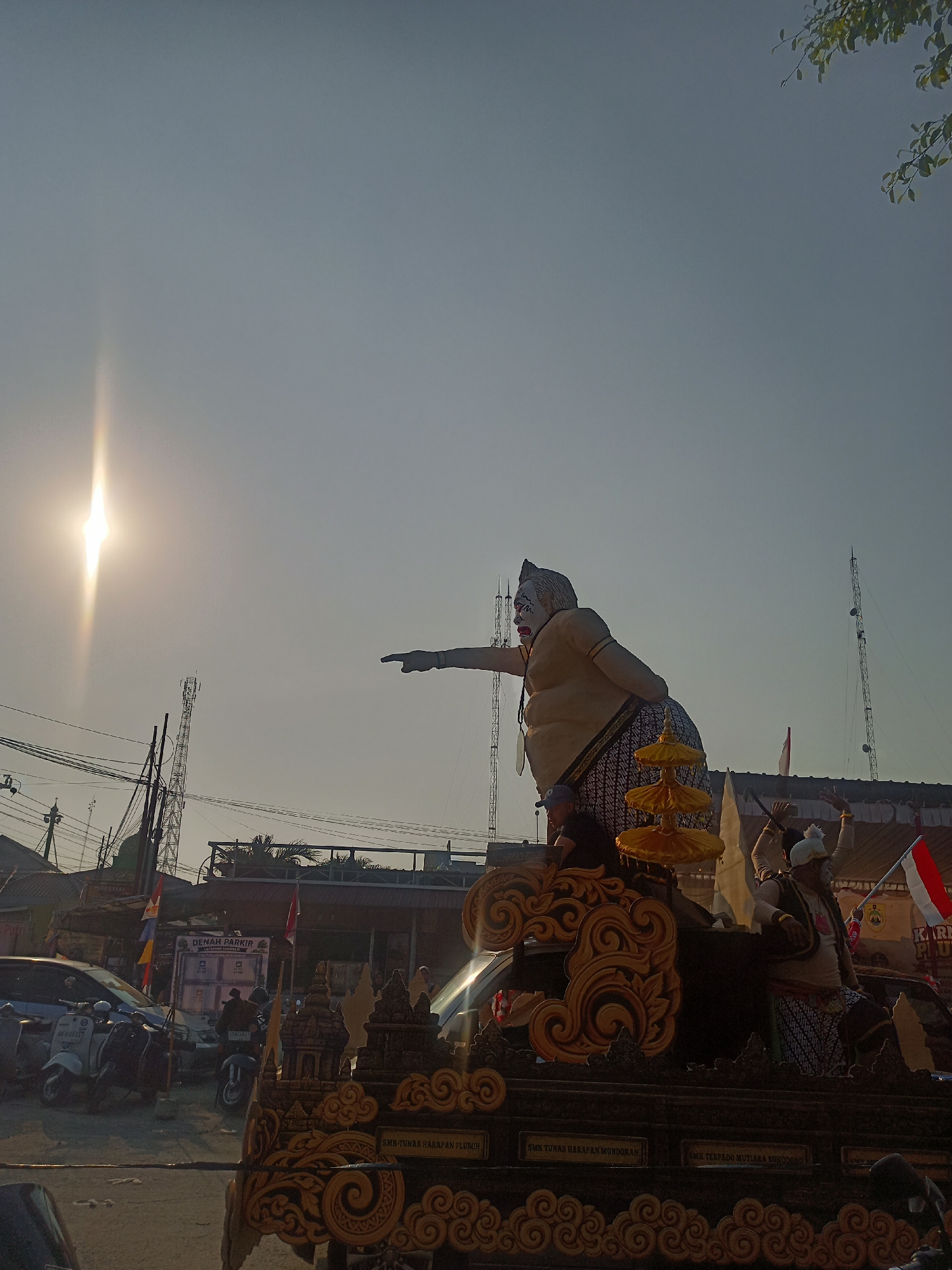 The image shows a large, colorful float with figures, likely part of a parade or festival, in the city of Ngrompongan, Indonesia. The sun is low in the sky, creating a bright lens flare on the left side of the image. On the float, a prominent, oversized figure with a pale complexion and a white face is dressed in traditional Indonesian attire, including a patterned sarong. This figure is pointing to the left. Next to it, a smaller figure in similar clothing is also depicted. The float itself is elaborately decorated with gold ornamentation and traditional architectural motifs. In the background, there are buildings, electrical poles, cell towers, and parked vehicles, including scooters and a car. The sky is clear and light blue. The scene appears to be outdoors in a somewhat urban setting.