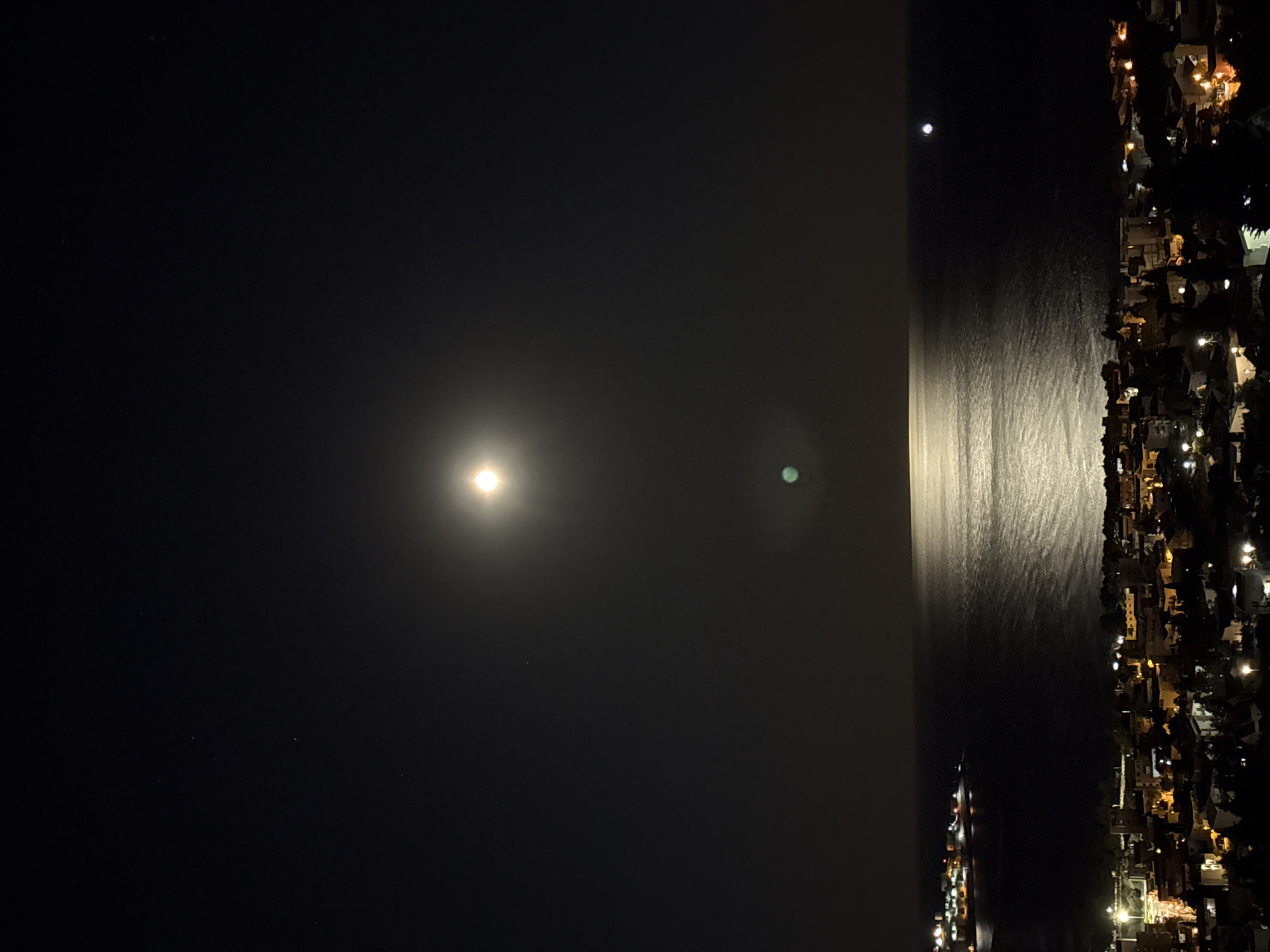 An aerial photograph captures the night scene in Šušanj, Montenegro. The full moon dominates the sky, with its light reflected on the still water below. The city lights from the buildings along the shore illuminate the coastline. The photograph does not show people or specific actions; instead, it captures the overall serene atmosphere of a coastal city at night.