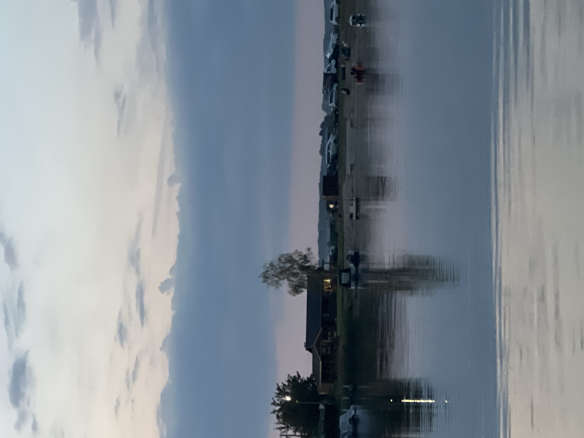 This is a photograph of a waterfront scene, likely during twilight, with a body of water in the foreground and a building complex and vehicles in the background. The sky is overcast with soft clouds. On the left side of the background, there is a building with a sloped roof, and several trees are visible, one of which is a tall, slender birch tree. In front of the building, on the water, are several covered boats. Further to the right, in the background, is a collection of vehicles, including cars, a truck, and what appear to be vans or SUVs, parked on what seems to be a sandy or gravel area. There are also a few small boats and what could be a pier or dock. The water reflects the sky and the structures on the shore. The scene is calm and quiet, suggesting it might be a recreational area or a residential waterfront.