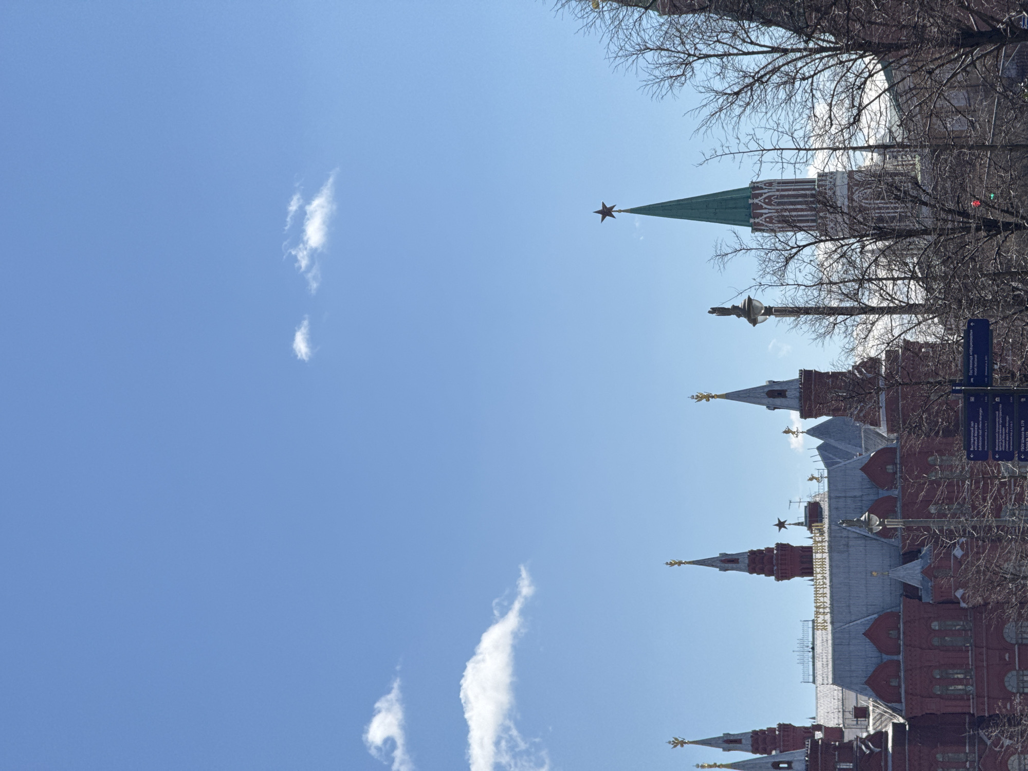 The image captures a view of the Moscow Kremlin in Moscow, Russia, against a bright blue sky with scattered white clouds. The Kremlin's red brick walls and towers are visible, with their distinctive green roofs and golden spires topped with stars. Bare tree branches are silhouetted against the sky in the upper right portion of the image. Street signs and lights are also visible on the right side of the image.