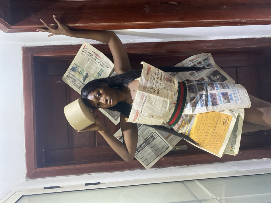 An indoor scene in Joma, Ghana, depicts a female subject posing in front of a dark wooden door adorned with newspaper pages. The subject, a woman with dark skin and long, straight dark hair, looks directly at the camera with a neutral expression. She is wearing a strapless dress fashioned from multiple newspaper pages, secured at the waist with a green and red striped band. Visible headlines on the dress include "E-Levy accrues GHC196.6m" and "Odumase farmers 160,000 cashew," alongside an "MTN" logo. Her left arm is raised, holding a light-brown cylindrical object approximately 15-20 cm in diameter above her head. Her right arm is bent and raised, with her fingers resting against the right side of the dark wooden doorframe. She wears a delicate necklace and a thin bracelet on her left wrist. The wooden door behind her features a metallic doorknob/handle on its left side and several scattered newspaper sheets affixed to its surface. A light-colored wall is visible to the left of the door.