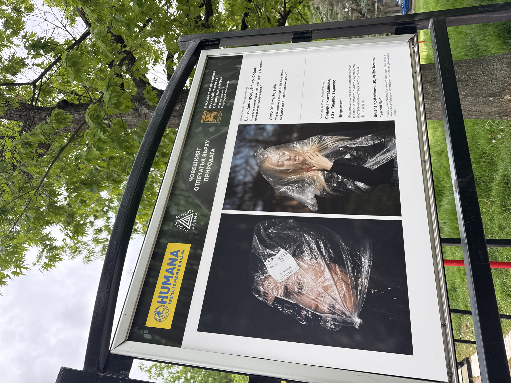Here's a detailed description of the image:
**Overview**
The image features a sign or display board outdoors. The sign is part of an exhibition titled "The Human Footprint on Nature," organized with the support of the Blagoevgrad Municipality. The location is presumably within the city of Blagoevgrad, Bulgaria, but there are no immediate landmarks or structures visible to confirm the location.
**Sign Details**
The sign is rectangular and framed in silver with a black metal support structure. The sign itself is printed on a white background with sections of black print and blue/gold accents.
There is a yellow logo in the upper left corner with the text "HUMANA PEOPLE TO PEOPLE BULGARIA". Adjacent to the logo is a triangle with a "School Dedal Varna" text.
**Photographs Displayed**
The sign features two photographs:
* **Left Photograph:** Shows a person's face inside a clear plastic bag. A price tag is attached to the bag. The photographer is Vanyo Dimitrov (39 years old, Sofia). The text below the photograph indicates the artist's concept, "The human as a product of his own time - packaged and assigned a market price."
* **Right Photograph:** This depicts a person (appears to be a woman) with a clear plastic bag covering their face/head. The photographer is Svilena Kostadinova (30 years old, Veliko Tarnovo). The accompanying text is "Second Skin" and explains that plastic today becomes like a second skin, a symbol of an era where the artificial replaces the living.
**Environment**
The sign is outdoors. Behind and above the sign, there is greenery, including tree branches and leaves.