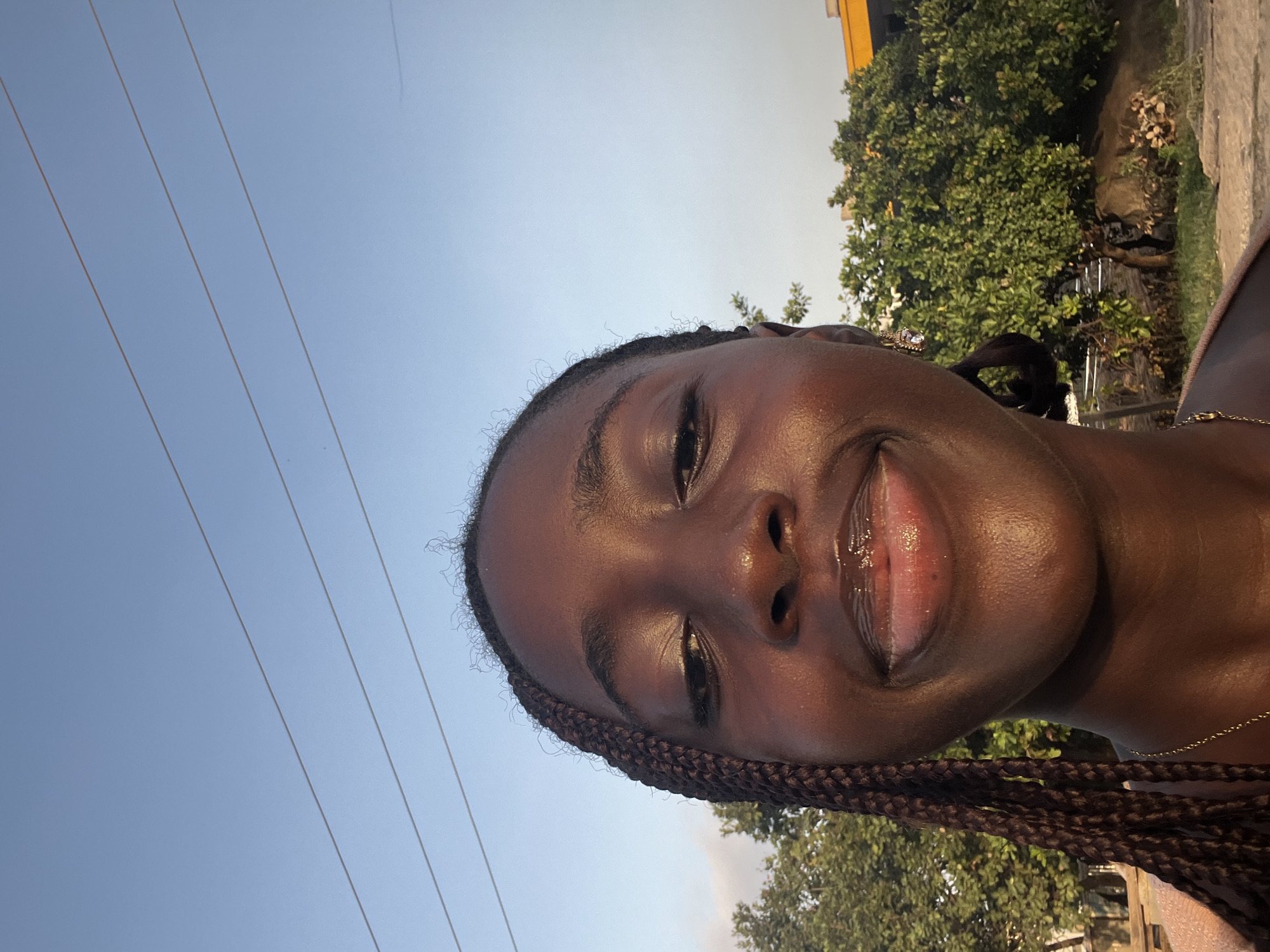 A young woman with dark skin and braided hair is taking a selfie outdoors. She is smiling with her lips slightly parted, revealing lip gloss. In the background, there are green trees, power lines against a blue sky, and a yellow building. The image appears to be taken in Karum, Nigeria.