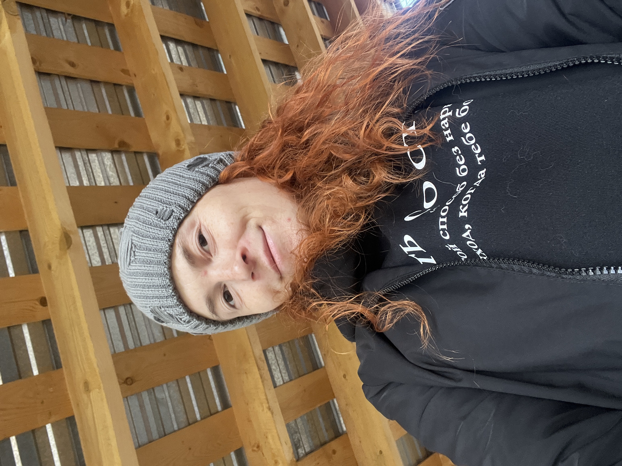 A person with red, curly hair wearing a gray beanie and a black jacket is positioned in front of a wooden lattice. The person is also wearing a black shirt with white Cyrillic text. The location appears to be outdoors. The city of Chaikovsky, Russia is not discernable from the image.