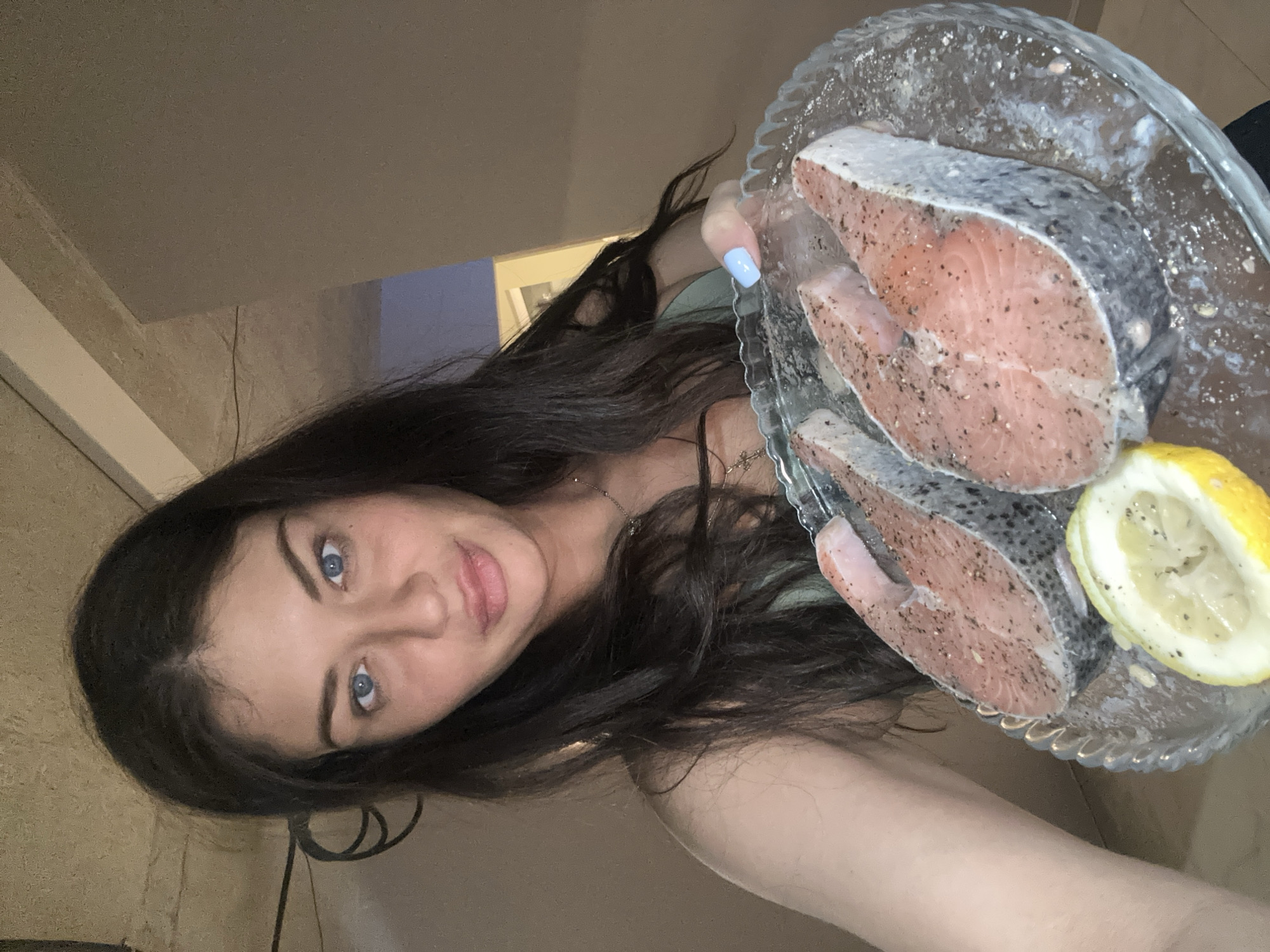 A young woman is captured in a vertical, close-up selfie, holding a clear glass plate containing three raw salmon or trout steaks, seasoned with black pepper. A bright yellow lemon half accompanies the fish on the plate, which features a decorative fluted edge. The woman has long, dark hair and striking blue eyes, looking directly at the camera with a soft, neutral expression hinting at a slight smile. She wears a light-colored top and a delicate necklace. Her index fingernail, visible as she grips the plate, is painted with a light blue polish.
The background suggests an indoor setting, likely a kitchen or dining area, with light-colored walls. A segment of a wall or doorway in the background appears to be a purplish-blue hue. The lighting is artificial, illuminating the scene. The image depicts a moment of food preparation or presentation. No specific time of day or external weather conditions are discernible from the indoor environment. There is no visible text. The scene is contextualized to Khimki, Russia.