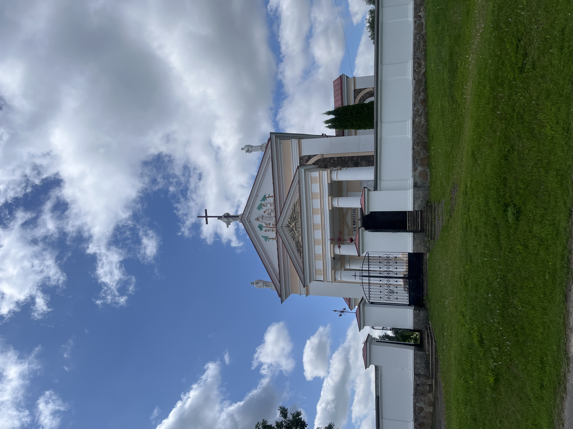 The image depicts the facade of the Church of the Transfiguration in Zhaludok, Belarus, under a partly cloudy blue sky. The church is a Neoclassical building with a prominent pediment topped with a cross and statues. The facade features a portico with columns and decorative elements. A white wall surrounds the church grounds, with a black wrought-iron gate in the foreground. A well-maintained green lawn stretches out in front of the wall. The overall scene is one of a serene, historical religious site bathed in daylight.