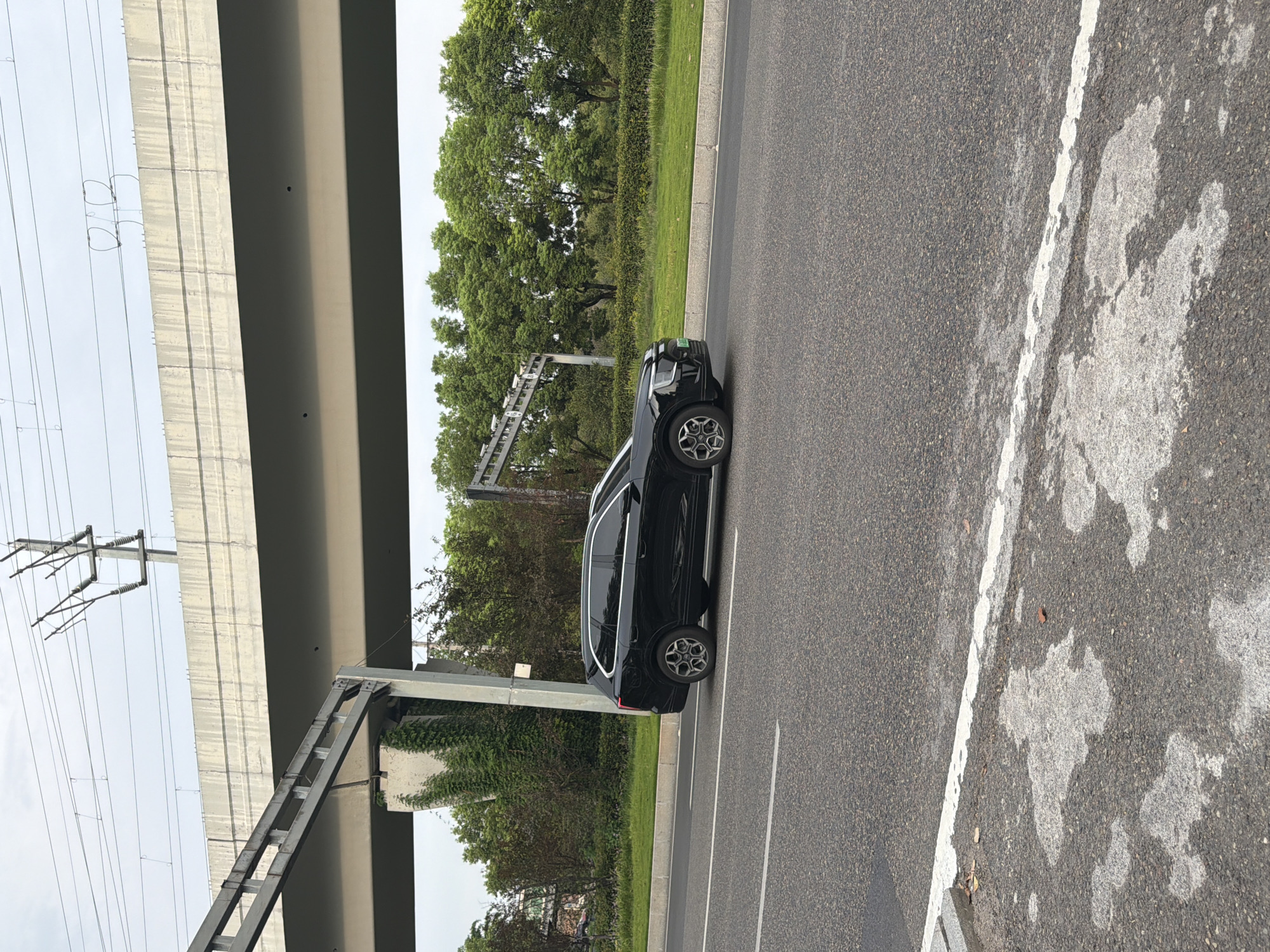 A black SUV is captured in profile, driving on a paved road under an elevated concrete overpass. The SUV has its headlights on and is positioned in the right lane. The overpass is supported by concrete pillars and a substantial concrete structure. Overhead, power lines and support structures are visible against a cloudy sky. The roadside features green grass and trees. The road surface is asphalt with white lane markings. The foreground of the image shows the road surface with white markings and patches of lighter colored material. The scene appears to be in a semi-urban or suburban environment, possibly along a major thoroughfare. The presence of overhead electrical infrastructure suggests proximity to transportation or industrial areas. The image seems to have been taken during daylight hours.