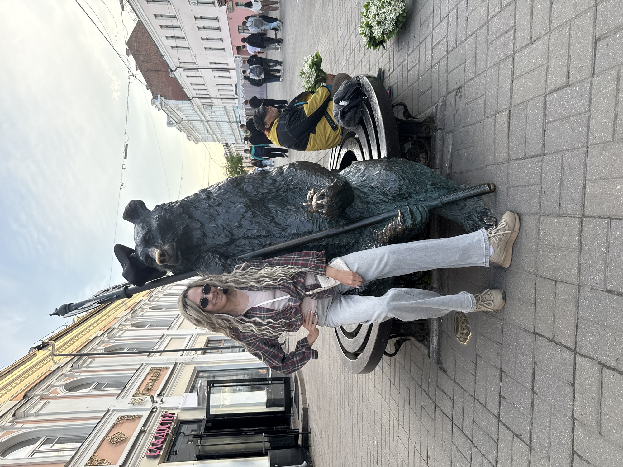 The image captures an urban street scene in Заволжье, Russia, featuring a young woman posing next to a large bear statue. The woman, with long blonde dreadlocks and sunglasses, is dressed in a red and black plaid shirt over a light top, light-wash jeans, and beige sneakers. She is smiling and looking towards the camera, holding a small white pouch.
To her right, a dark-colored statue of a bear sits on a decorative bench, holding a staff. A smaller, lighter-colored animal figure rests on the bear's left leg. In the background, another person wearing a yellow and black jacket is seated on the bench, holding a bouquet of white flowers. Several other pedestrians are visible walking along the grey-tiled sidewalk.
The street is lined with multi-story buildings, displaying varying architectural styles with numerous windows and decorative elements. The sky appears bright and partly cloudy. On one of the buildings in the background, a sign with Cyrillic text is visible, clearly showing "СЛАДОСТИ" (SLADOSTI), meaning "Sweets" or "Confectionery". The scene depicts a casual daytime moment, likely in a pedestrian area of the city.