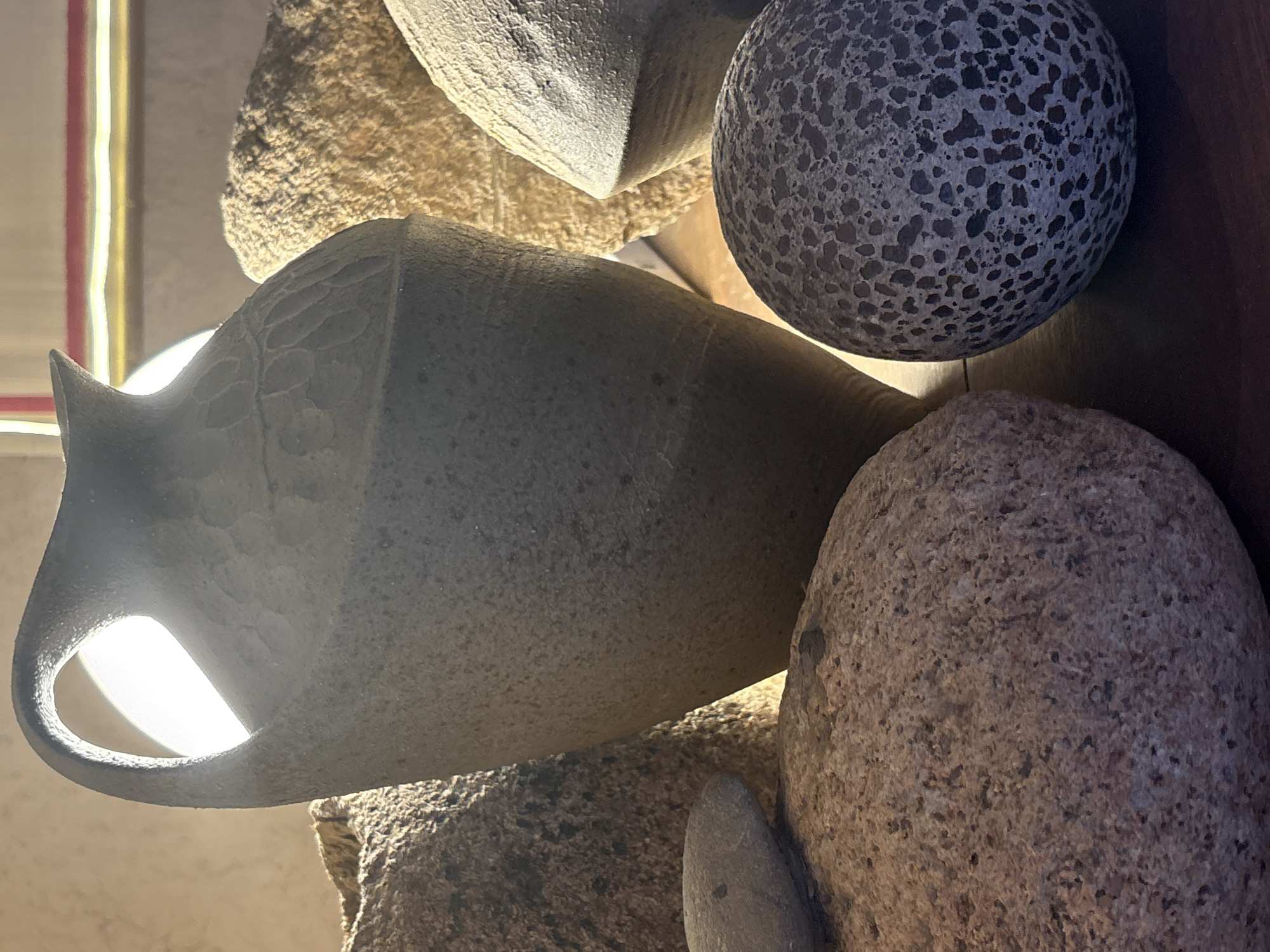 The image depicts a still life arrangement of textured objects, likely decorative, with a strong light source illuminating them from behind. The arrangement includes a light gray pitcher-like object with a handle, positioned prominently. Adjacent to the pitcher are several stones or rocks of varying textures and sizes, including one that is speckled with a dark gray color, and a larger brownish-gray rock. The items are arranged against an out-of-focus background. The exact location of the subject is not known.