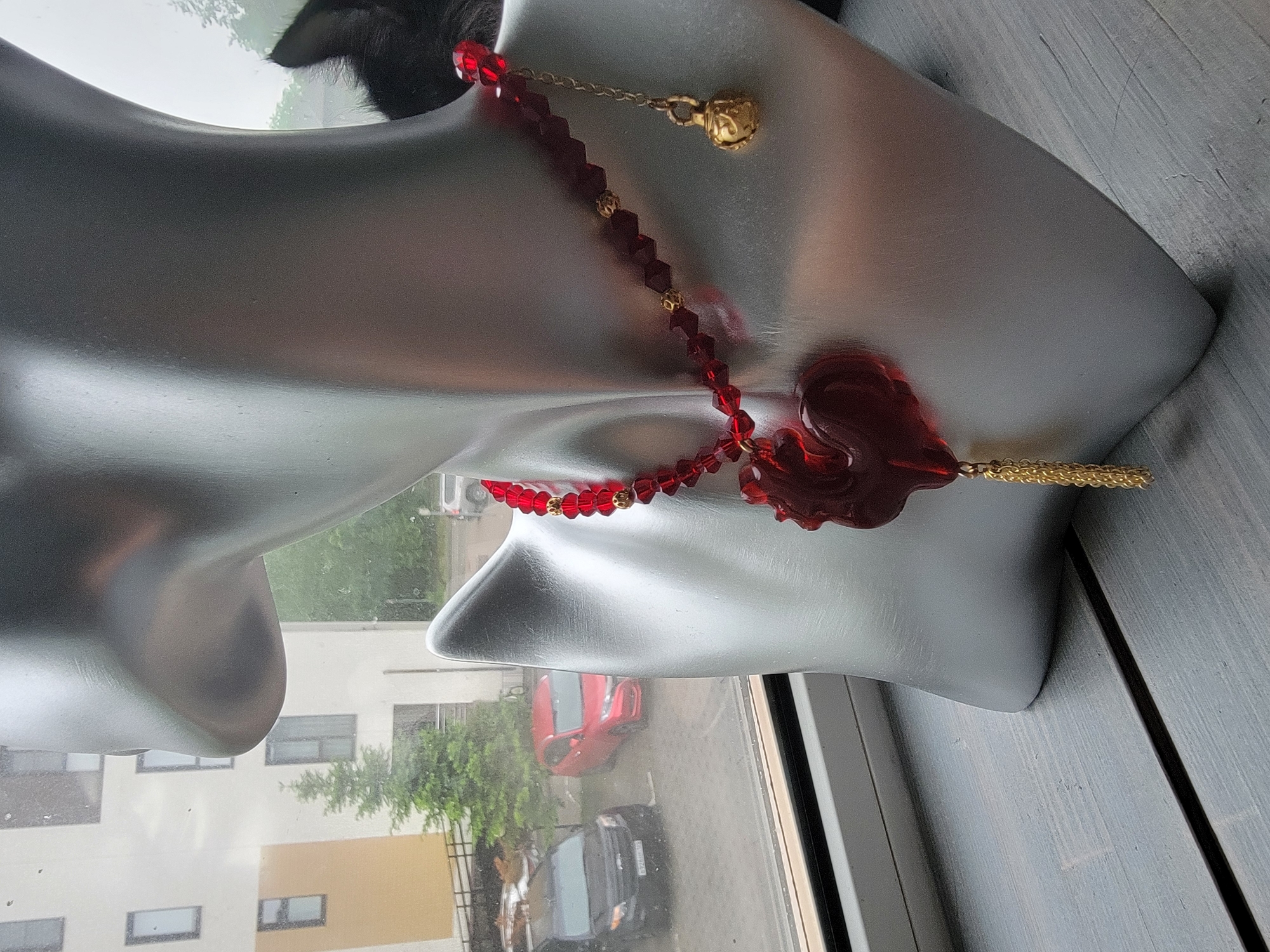 A close-up, eye-level shot depicts a gray mannequin head wearing a red bead necklace, positioned near a window. The necklace features a red, heart-shaped pendant with a gold tassel. Outside the window, a cityscape is visible, including a building and two cars: one red and one black. The scene appears to be lit by natural light. The setting is the city of Unknown, Unknown.