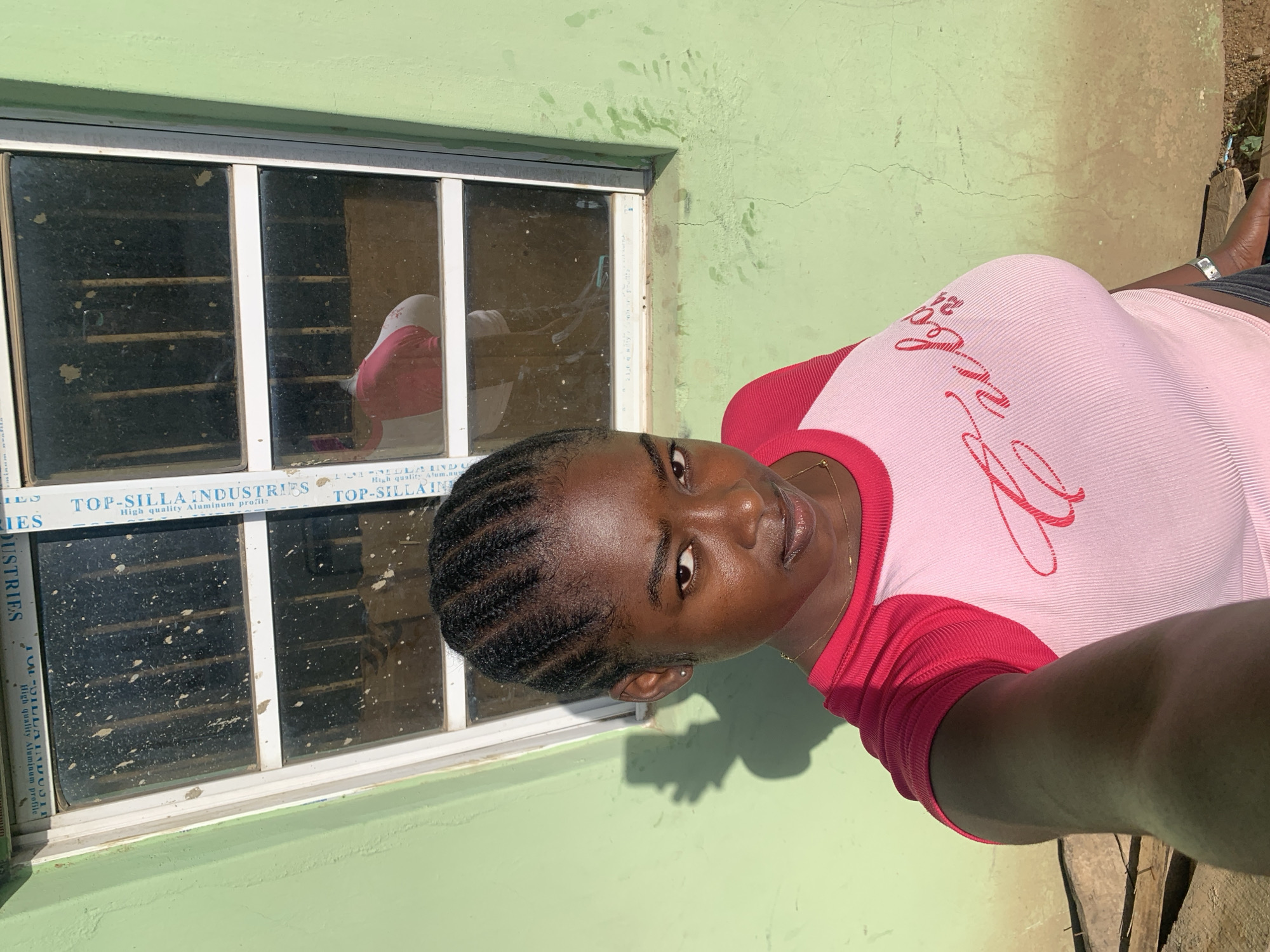 A young woman stands in front of a window. The window is framed with "TOP-SILLA INDUSTRIES" Aluminum profile. She is wearing a light pink shirt with red accents and her hair is braided. The building is painted a light green color. It is in Kabong, Nigeria.