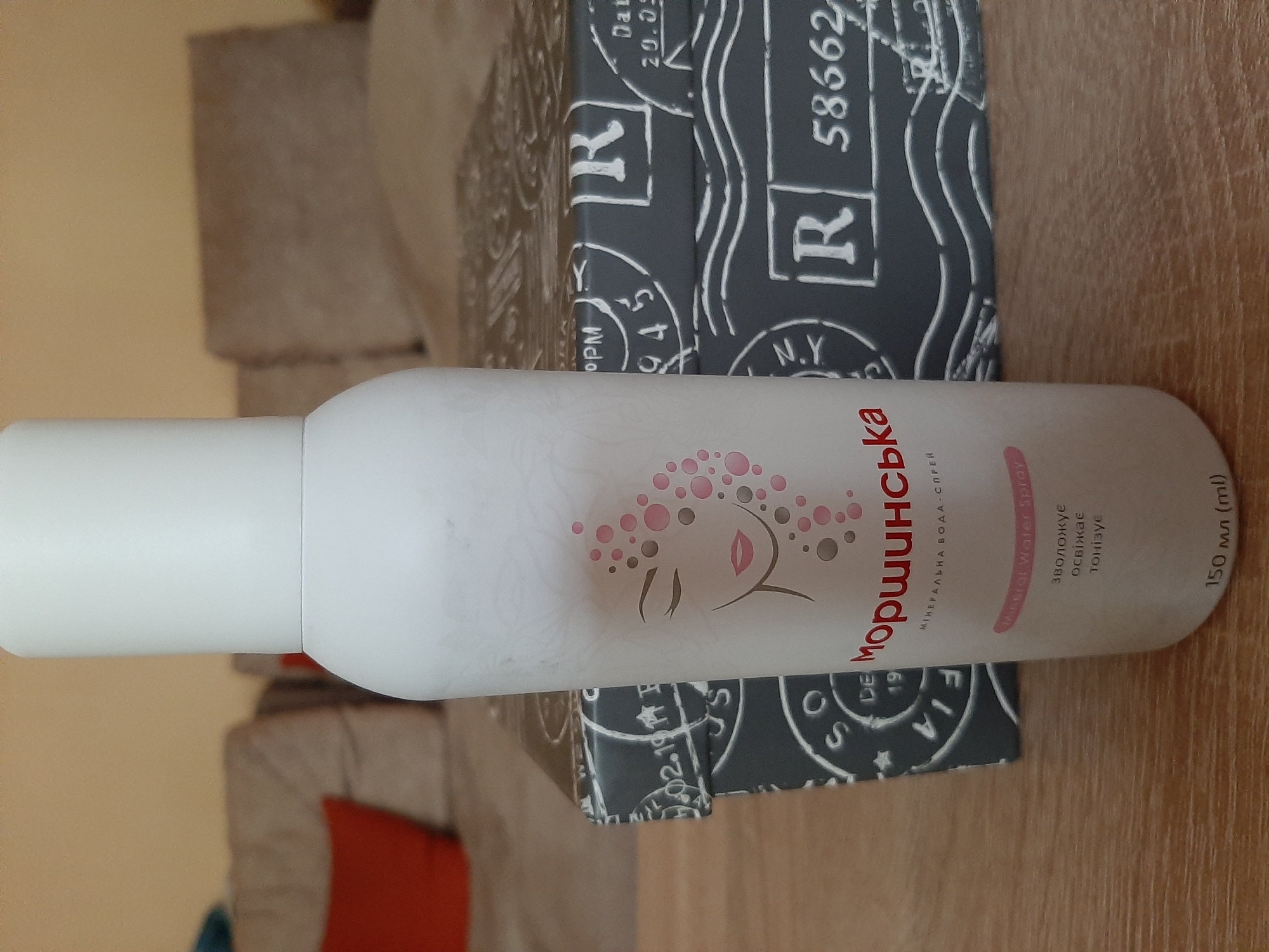The image shows a white aerosol can with pink and gray circular patterns and a stylized face on it, placed on top of a patterned box. The box has a design resembling postage stamps with the letters "R", "N.Y.", and numbers like "58662". The can appears to be a beauty product, possibly a mineral water spray, as indicated by the text on it. The text on the can is in Ukrainian and English. In Ukrainian, it reads "Моршинська" (Morshynska), "МІНЕРАЛЬНА ВОДА-СПРЕЙ" (Mineral Water Spray), and "зволожує освіжає тонізує" (moisturizes, refreshes, tones). The English text also says "Mineral Water Spray". The volume is stated as "150 мл (ml)". The patterned box has text like "Dat 20.05", "02.19", and "PM". The overall scene is indoors, with a wooden surface visible in the foreground and a blurred background suggesting furniture like a sofa with orange cushions. The lighting appears to be artificial and the setting is casual. The image is oriented vertically, with the can angled slightly upwards.