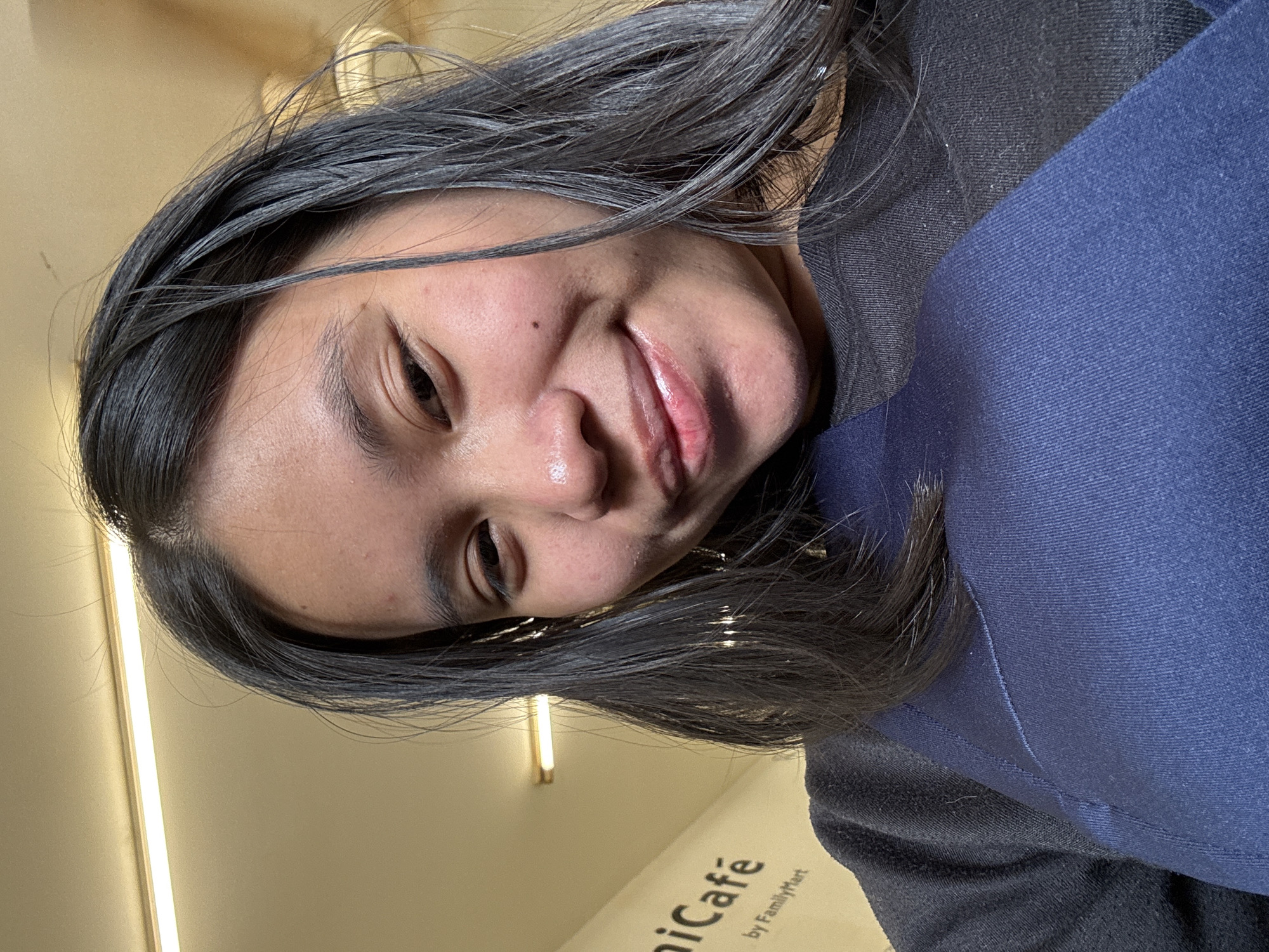The image shows a close-up, low-angle selfie of a young woman with long, dark hair and fair skin. She is looking towards the camera with a slight smile and her mouth slightly open. Her head is tilted, and her hair is falling loosely around her face. She is wearing a dark-colored top, possibly a sweatshirt, with a darker blue fabric overlaying the front.
In the background, to the left, a white sign with black lettering is visible. The visible text reads "nicáfe" and below it, "by FamilyMart". This suggests the location is likely a FamilyMart convenience store that offers a "nicáfe" service. FamilyMart is a prominent convenience store chain in Indonesia, and this particular establishment is located in East Jakarta, Indonesia. The ceiling above appears to be illuminated by strip lighting, common in retail environments.