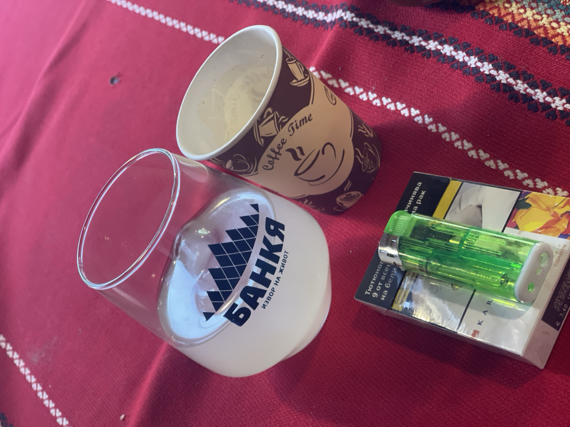 A glass of white liquid with ice, a disposable coffee cup, and a pack of cigarettes with a green lighter are arranged on a red tablecloth. The glass has black text and a mountain logo. The coffee cup has the words "Coffee Time" printed on it. The tablecloth has a white embroidered border.
