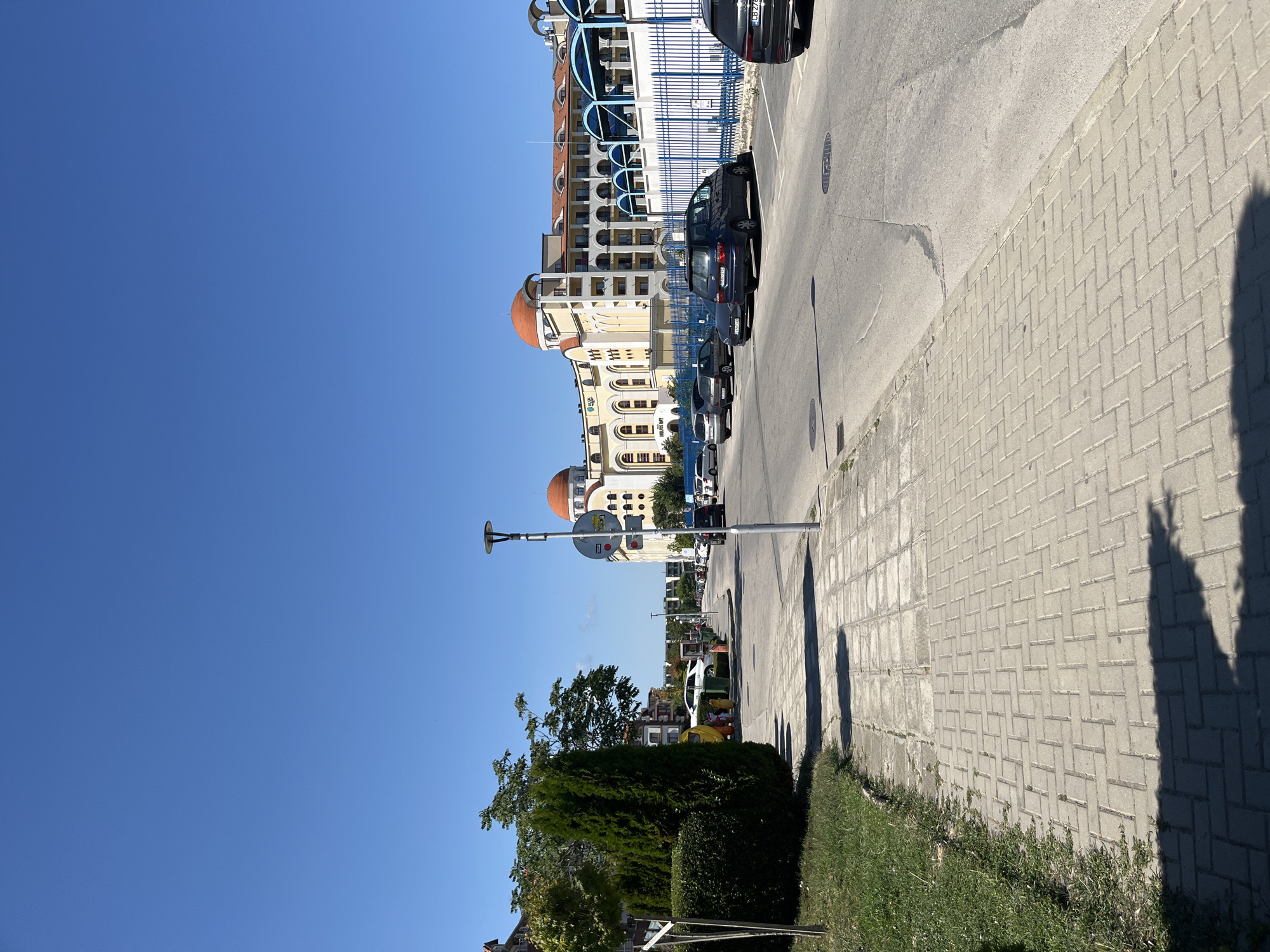 The image shows a street in Obzor, Bulgaria. A paved sidewalk runs along the left side of the image, bordered by a patch of grass. Across the street, several cars are parked. In the background, a large, light-colored building with orange domes dominates the scene. A blue fence runs in front of the building. The sky is clear and blue.