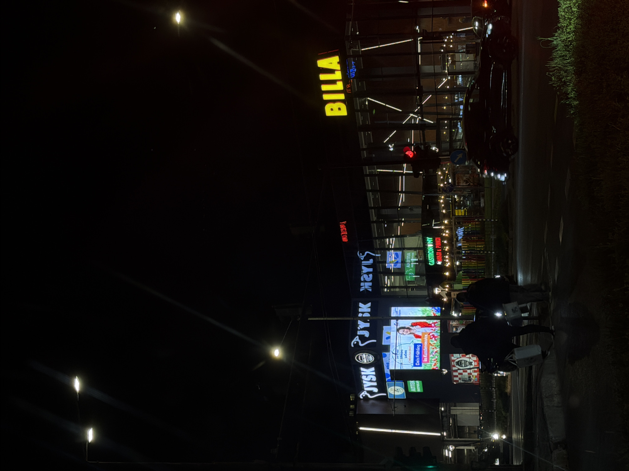 The image captures a night scene in Varna, Bulgaria, showing a street with illuminated commercial establishments. Notable buildings include Billa and JYSK stores, both marked with prominent signage. People are seen crossing the street, their figures silhouetted against the light from the stores. The environment is a mix of urban elements, including streetlights, roads, and sidewalks, as well as some greenery on the right side of the frame.