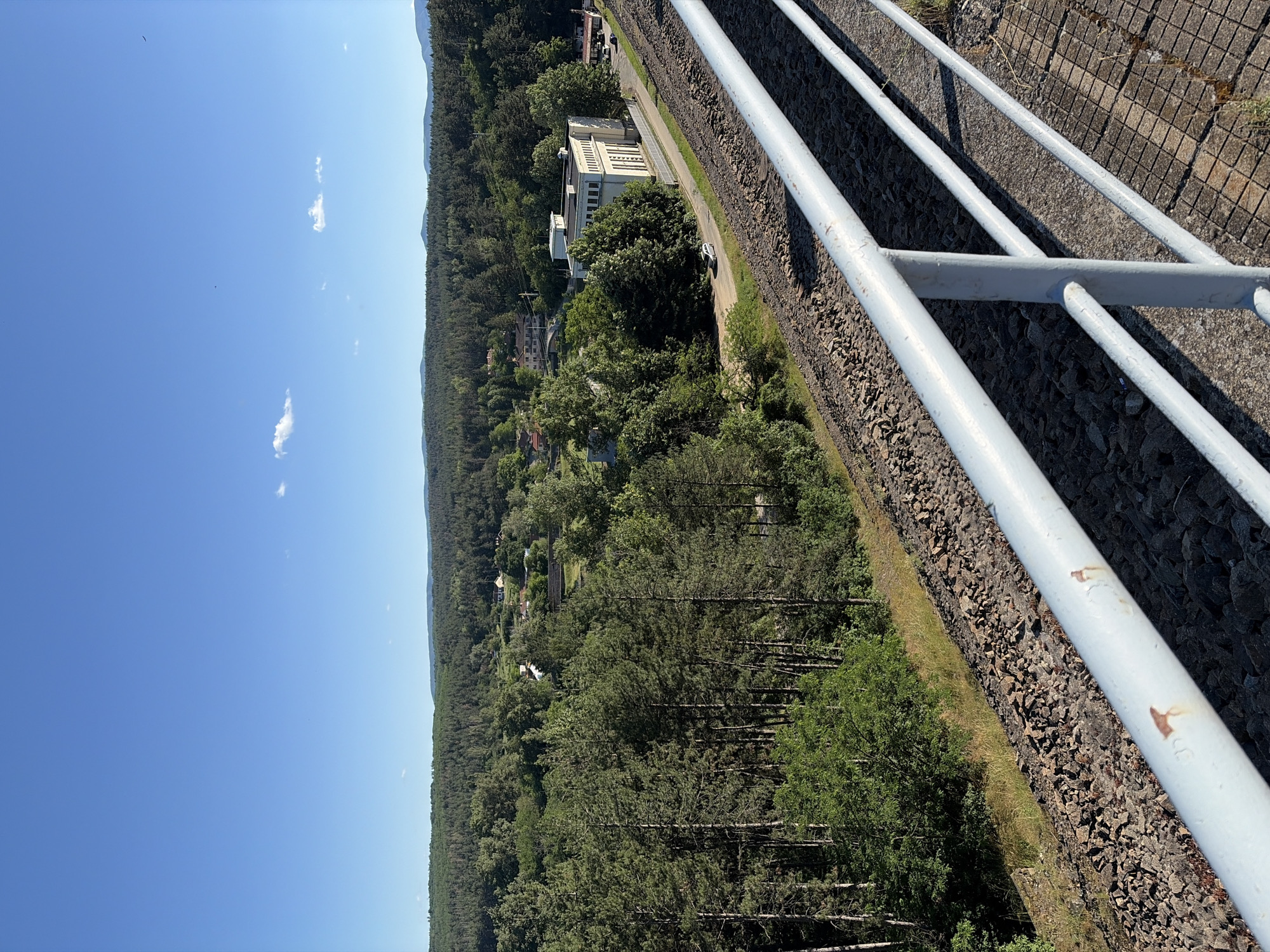 The image shows a wide-angle view from a high vantage point overlooking a landscape. In the foreground, a light-colored metal railing with multiple horizontal bars runs diagonally across the frame, obscuring a portion of the scene below. The railing is mounted on a concrete surface with a grid pattern. Below the railing, a steep embankment covered in dark, jagged rocks slopes down. Beyond the embankment, a railway track is visible, consisting of gravel ballast and parallel metal rails. To the left of the tracks, a dense forest of tall, green pine trees dominates the mid-ground, with some deciduous trees mixed in.
Further into the scene, nestled amongst the trees, are scattered buildings, including houses with red roofs and a larger, pale-colored, multi-story building with a flat roof, resembling a power station or industrial facility. A paved road runs alongside this building, with a few parked cars. The background features rolling hills covered in a continuous expanse of forest, stretching to the horizon under a clear blue sky with a few wispy white clouds. The overall impression is one of a rural or semi-industrial area surrounded by nature. The location is identified as Koprinka, Bulgaria.