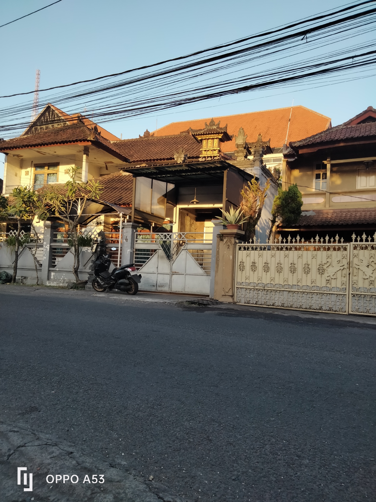 A motorcycle is parked on a street in front of a house with traditional Balinese architecture. The house has a red tile roof and intricate decorative elements. The street is paved and appears to be in a residential area. The sky is clear and blue, indicating a sunny day. The image is taken from a low angle, looking up at the house and street. A cell tower is visible in the background. The scene is likely located in the city of Denpasar, Indonesia, given the architectural style.