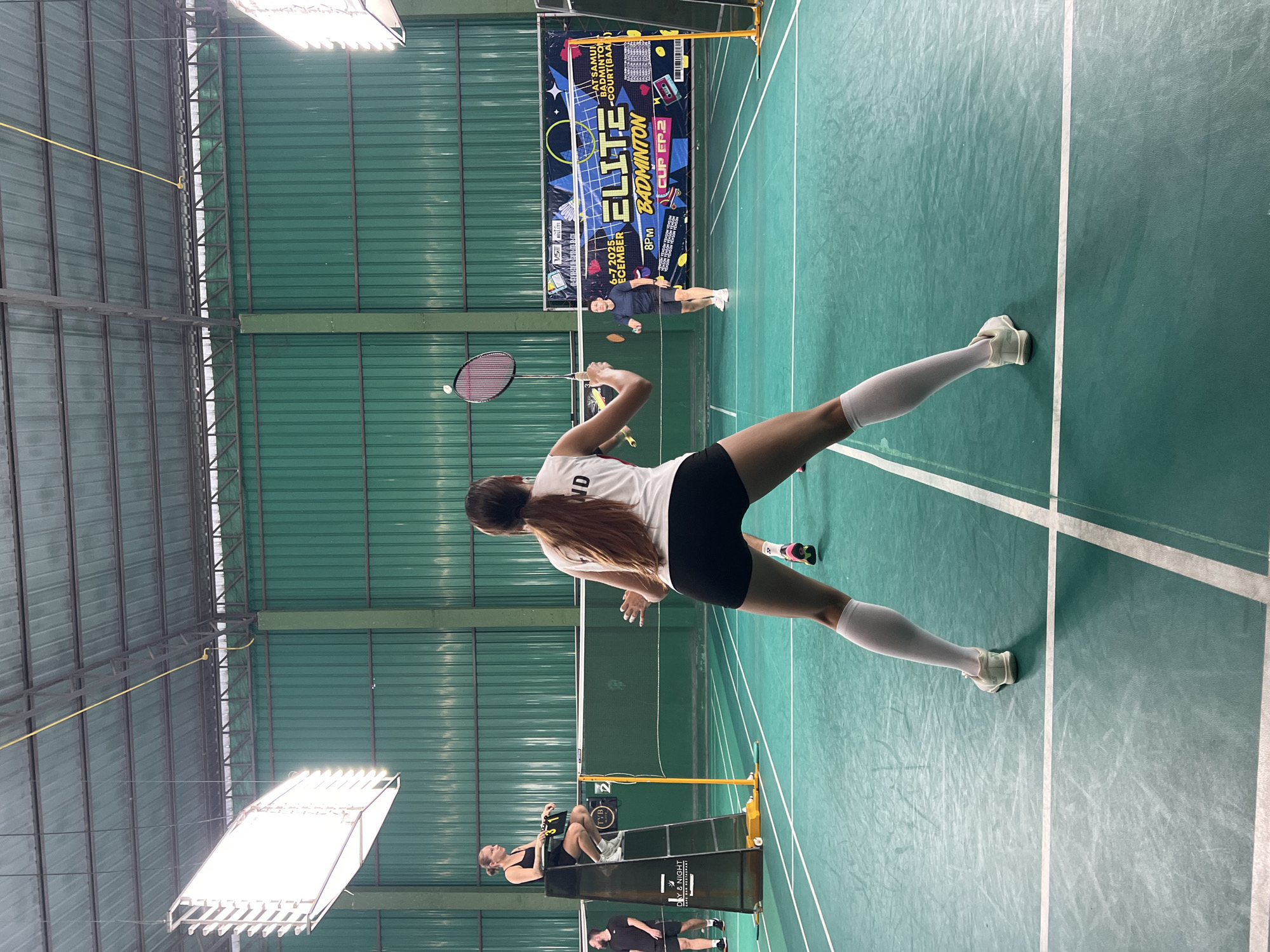 The image depicts an indoor badminton court, likely in the vicinity of Ban Bang Rak, Thailand, based on the presence of a banner indicating "ELITE BADMINTON COURT (BAA)". The court has a green playing surface with white lines. In the foreground, a woman in athletic attire, including white knee-high socks, is positioned in a ready stance, holding a badminton racket. She is facing away from the camera, with her back to the viewer, and appears to be mid-game. Another player, a man in a dark t-shirt and shorts, is visible on the opposite side of the net, in motion. Further in the background, a female umpire is seated on a high chair on the left side of the court. The court is illuminated by bright overhead lights mounted on the ceiling. A large banner with text and graphics, likely related to a badminton tournament, is displayed on the back wall. The overall environment suggests a professional or semi-professional badminton facility.