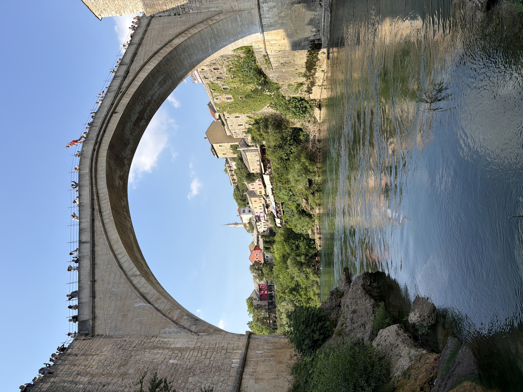 The image depicts a view from the Neretva River looking towards the iconic Stari Most (Old Bridge) in Mostar, Bosnia and Herzegovina. The bridge, a UNESCO World Heritage site, is constructed of stone with a large arch spanning the river. On top of the bridge, a crowd of spectators is gathered, observing an event. One individual, dressed in red shorts, is positioned at the edge of the bridge, appearing to be preparing to jump. The river flows calmly below, reflecting the blue sky and surrounding buildings. The riverbanks are lined with lush green vegetation and rocky outcrops. On the far side of the river, the historic old town of Mostar is visible, with its characteristic Ottoman-era houses painted in various colors, nestled on the hillside. A minaret of a mosque can be seen rising above the rooftops, indicating the city's rich cultural heritage. The scene is bathed in the warm light of a sunny day.