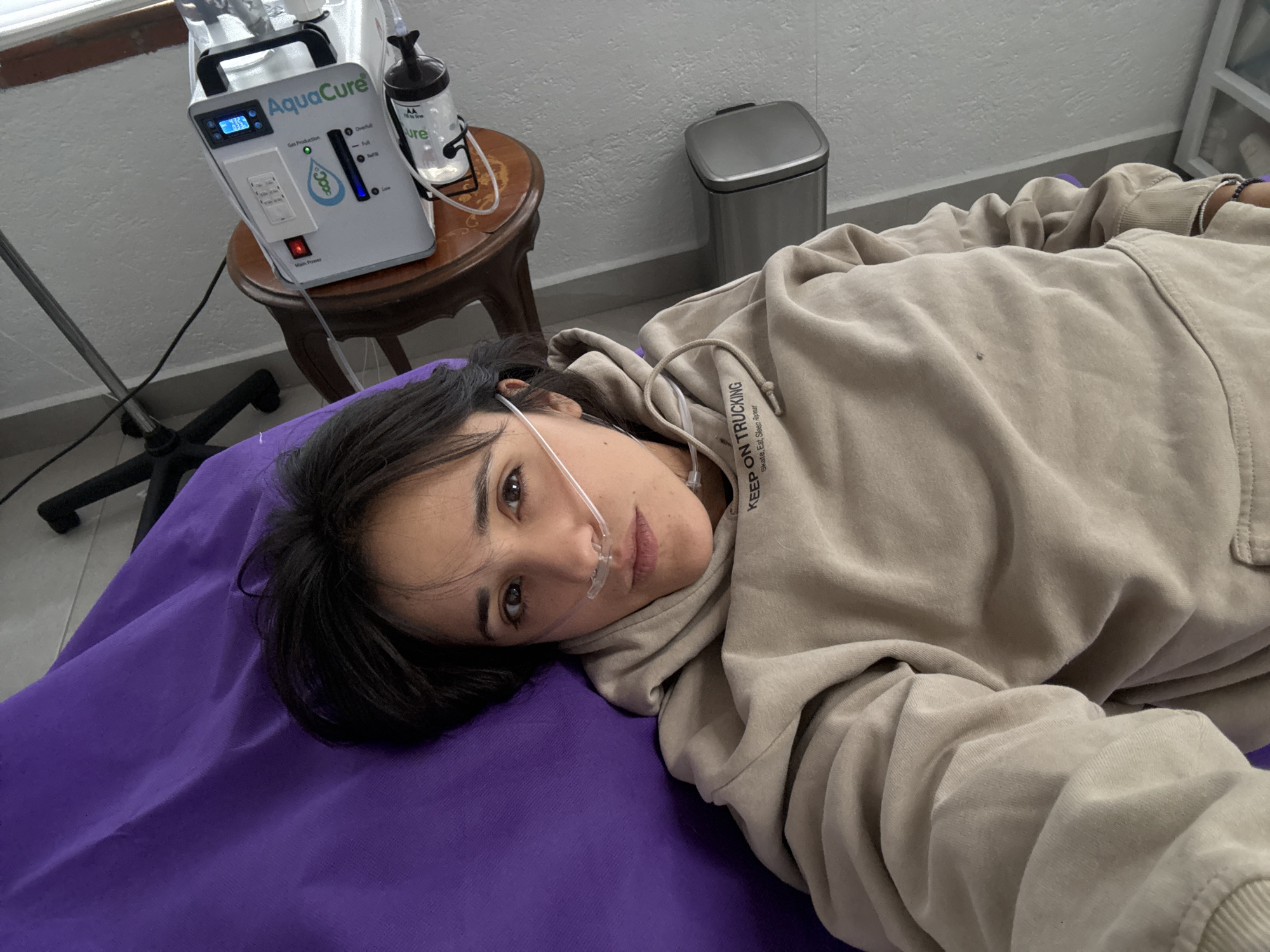 A woman lies on a purple surface, appearing to be a medical or treatment table. She is wearing a beige hoodie with the text "KEEP ON TRUCKING" printed on it. A nasal cannula is in place, suggesting she is receiving some form of oxygen therapy.
Next to her on a small wooden table is a device labeled "AquaCure", a machine used for producing hydrogen and oxygen.
In the background, a stainless steel trash can can be seen. The room appears to be a treatment room. The scene is likely set in a medical or wellness facility, possibly in Mexico City, Mexico.