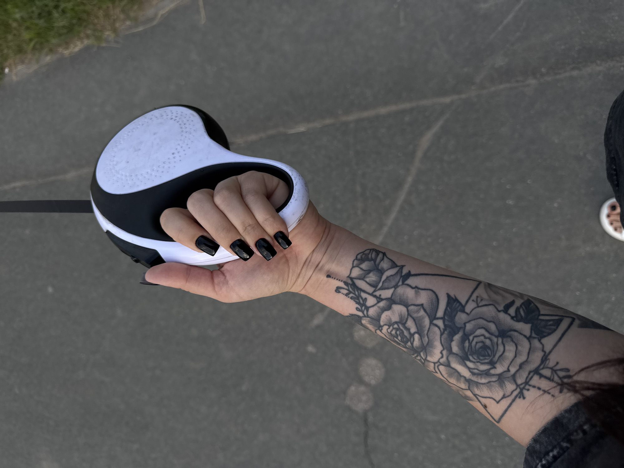A person's hand, with black painted fingernails, holds a black and white retractable dog leash. The person's left arm, visible from the wrist down, is adorned with a black ink tattoo of roses and a geometric design. The background consists of a grey asphalt surface with faint circular markings and a strip of green grass along the right edge. The scene appears to be outdoors, potentially in an urban or suburban setting, as indicated by the asphalt and grass.