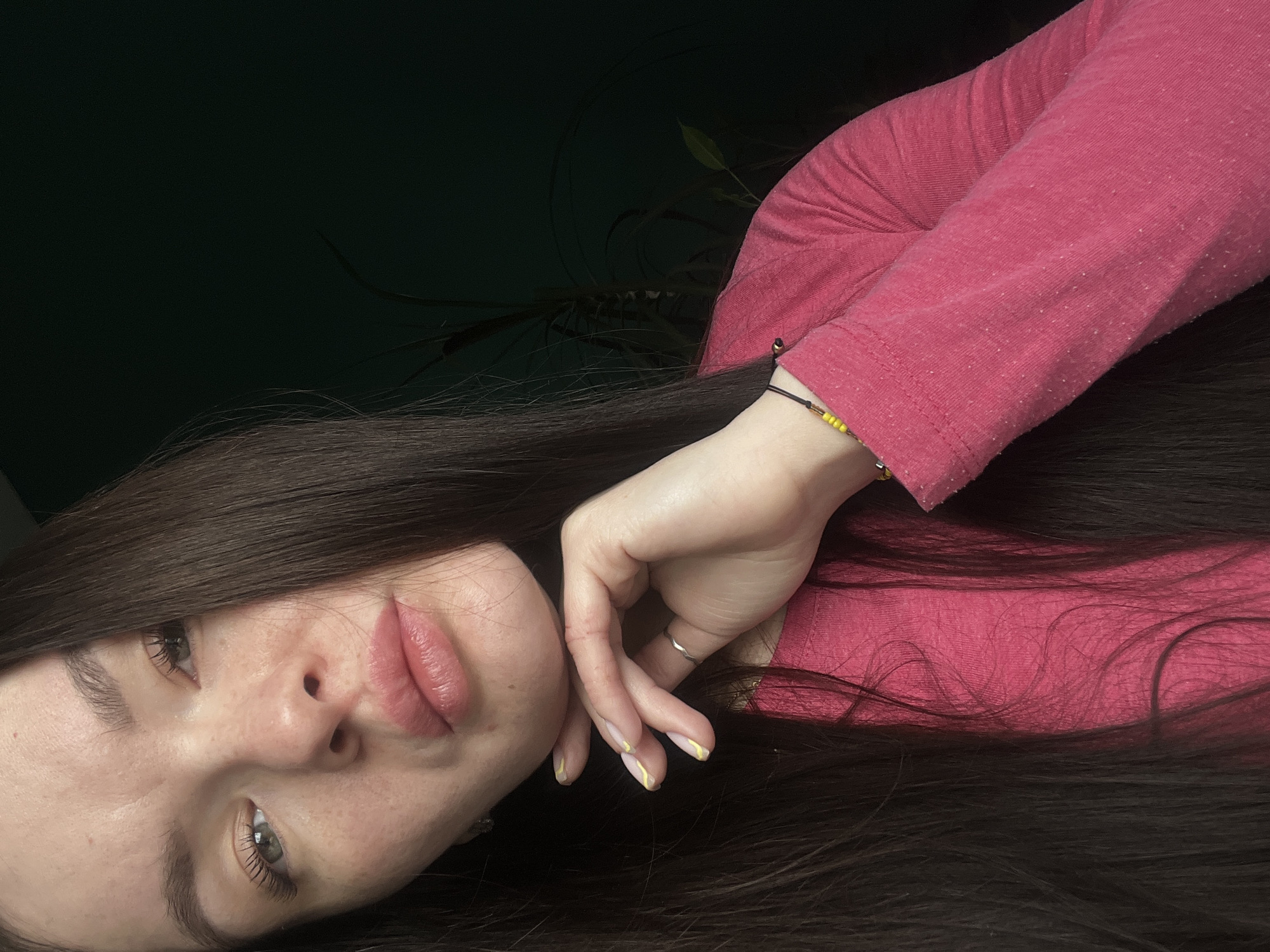 A woman with long, dark brown hair is lying down. She is wearing a pink long-sleeved shirt. Her left hand is resting on her face, and she is wearing a bracelet with yellow beads on her left wrist and a ring on her left ring finger. Her nails are painted with a yellow design. The background is dark and appears to be a wall and a potted plant with green leaves. There is no information available in the image that indicates the city of Kazan, Russia.