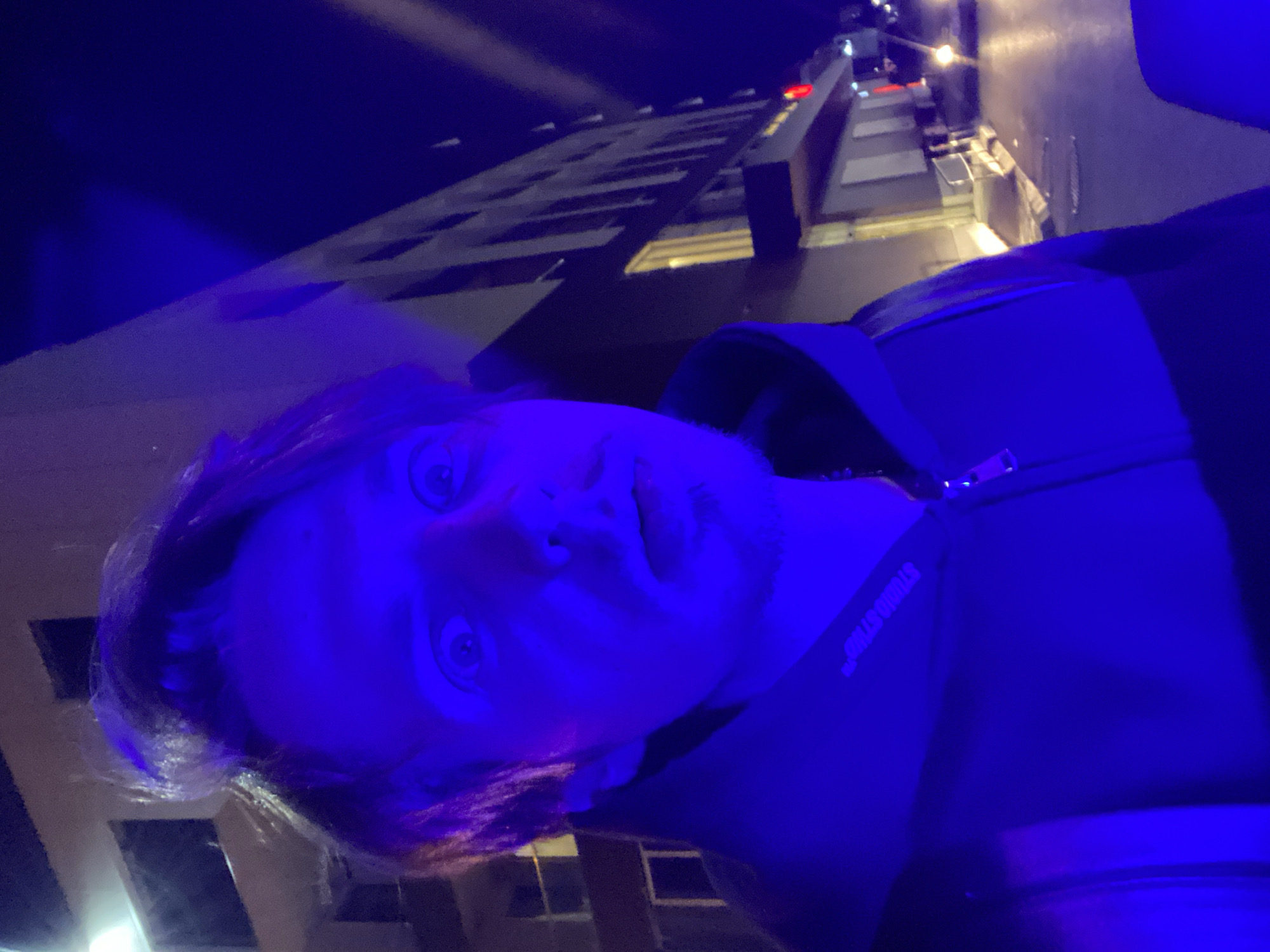 A selfie of a fair-skinned male with light brown hair and beard is bathed in intense blue light. He is wearing a dark jacket with the word "STUDIO STUFF" visible on the collar. His eyes are wide and he is looking directly at the camera. The background features a high-rise building with lit windows, typical of an urban environment. A street scene is partially visible, illuminated by streetlights. The perspective suggests the photo was taken at a low angle. The location is not discernible.