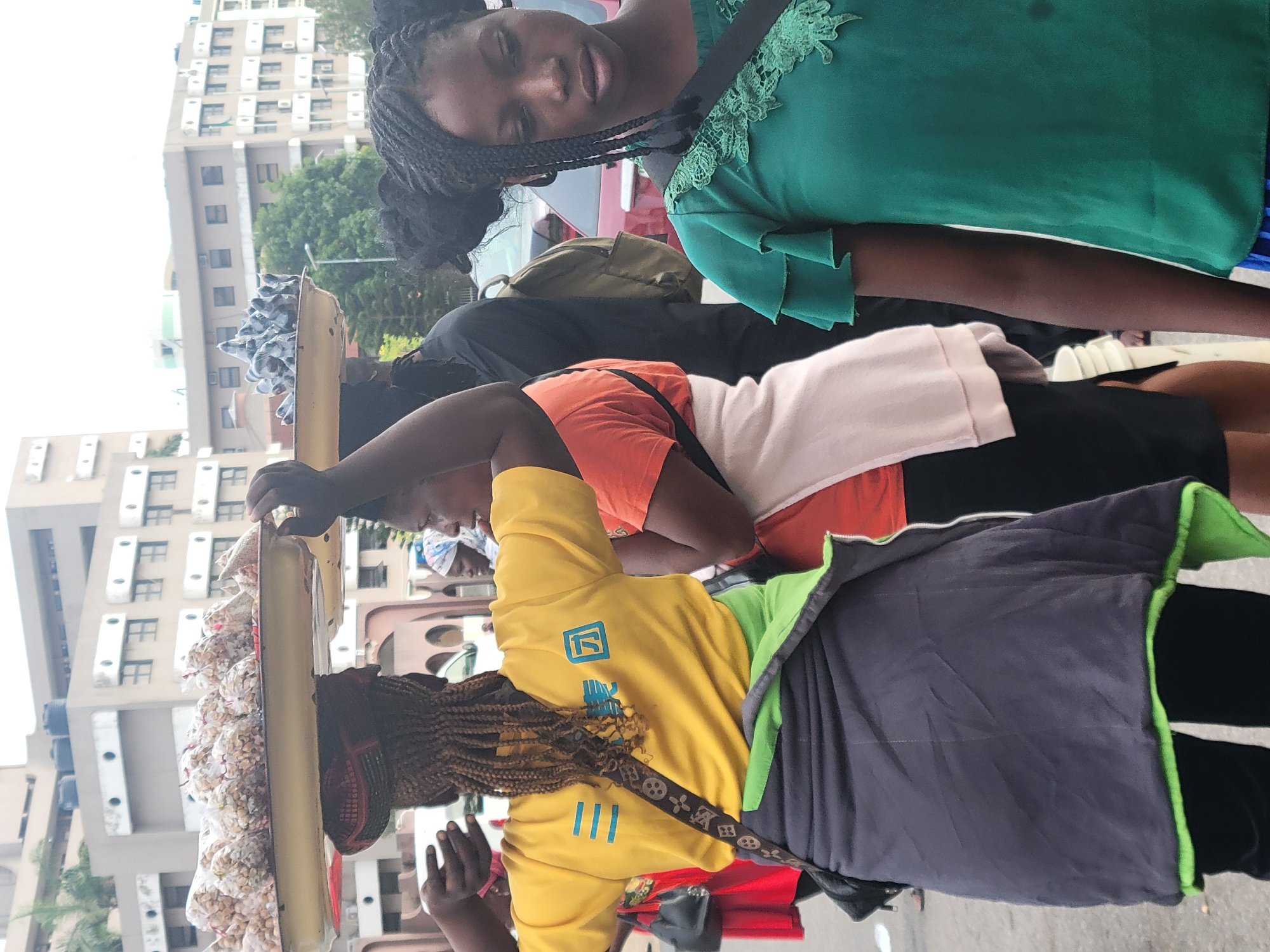 The image captures a street scene in Abuja, Nigeria, featuring several individuals. A woman in a yellow top with blue markings and an LV-patterned crossbody bag is carrying a tray of packaged snacks or food items on her head. Another woman wearing an orange top and a black skirt is behind her. The top part of a person in a green top with lace details is visible in the upper part of the frame. Background buildings with uniform window arrangements suggest an urban environment. The scene appears to be a bustling street.