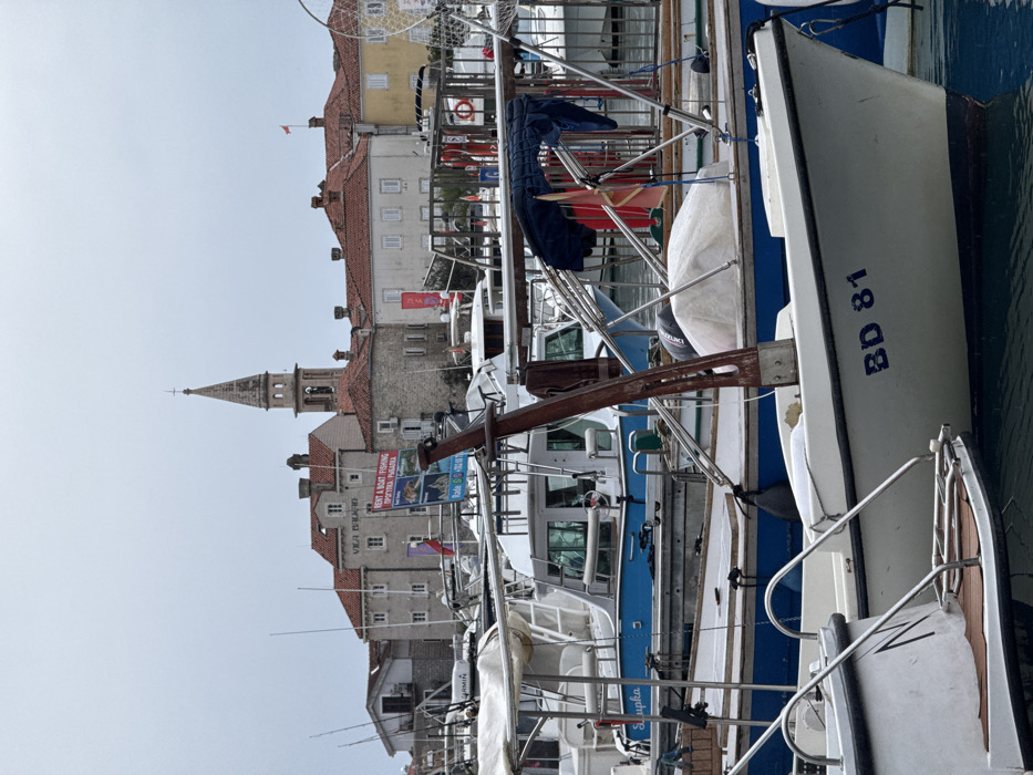 The image depicts a marina scene in Budva, Montenegro, with the historic Old Town (Stari Grad) visible in the background under an overcast sky.
In the foreground, numerous boats are moored in calm water. A white boat in the lower right foreground displays "BD 81" in blue lettering on its bow. To its left, another white boat shows part of a stainless steel boarding ladder. Behind these, a blue-hulled boat with a white cabin and the partial name "Slupka" is visible, featuring a "GARMIN" decal on an antenna mast. Various other boats, mostly white, are present, exhibiting details like fishing rods, metal railings, a red life preserver, and a Suzuki outboard motor. A prominent, rust-brown, crane-like structure rises from a boat in the mid-ground. Covered objects under white tarps are present on several decks, along with blue tarps and canopies. A portion of a fishing net is visible on the far right.
The background is dominated by the architecture of Budva Old Town. A tall, stone bell tower with a spire, characteristic of the Church of St. John the Baptist, stands centrally. Buildings with red-tiled roofs and light-colored stone or stucco facades are arranged around the tower. One building to the left of the tower features the inscription "VILA BUDVA." A multi-panel blue and white sign, prominently displayed among the boats, advertises "RENT A BOAT FISHING" in English and Cyrillic, with accompanying photographs and contact information "Radu S. +382 (0)67." Small flags, including red and white ones, are visible on some buildings and boat masts. The sky is uniformly light gray, indicating heavy cloud cover. No individuals are discernible in the scene.