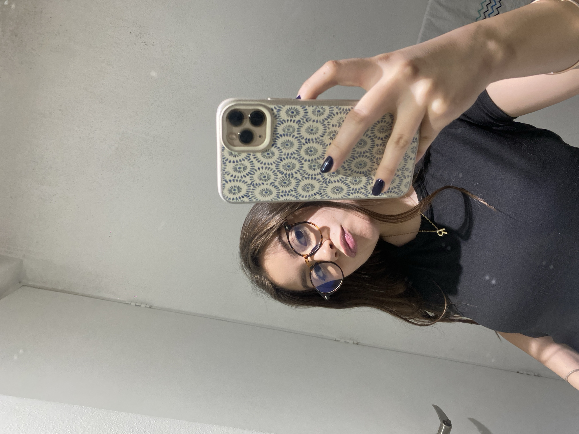 A woman is taking a selfie in an indoor environment. She is wearing glasses, a black shirt, and a necklace with a pendant. The phone she is using has a patterned case. The background appears to be a plain wall. The location is unknown.