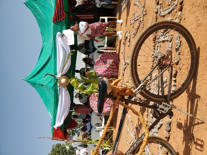 The scene depicts an outdoor event in Bar Gada, Nigeria. A black mountain bicycle with "GIANT" branding stands in the foreground on reddish-brown dirt, propped by a kickstand. The bicycle is decorated with a thick, braided tan rope wrapped around its frame and handlebars. Attached to the front of the bicycle are a bundle of green leaves, a small woven straw hat, and a curved, horn-like object.
In the midground, a large tent with a green tarp roof provides shade. The tent's front is decorated with white and red fabric drapes, along with green fronds and a small woven straw hat. Beneath the tent, numerous individuals are seated on white plastic chairs; some chairs have red and white checkered fabric covers. A young woman, wearing a white t-shirt, a red and white checkered skirt, and white sneakers, is standing in front of the tent, mid-movement, holding a white sign in her left hand. Other attendees in various attire are visible seated under the tent.
The dirt ground around the bicycle and extending into the midground is adorned with patterns, including partial letters and an anchor-like symbol, formed from light-colored stones or debris. The background consists of a clear blue sky and sparse trees on the left. The scene is illuminated by bright daylight.