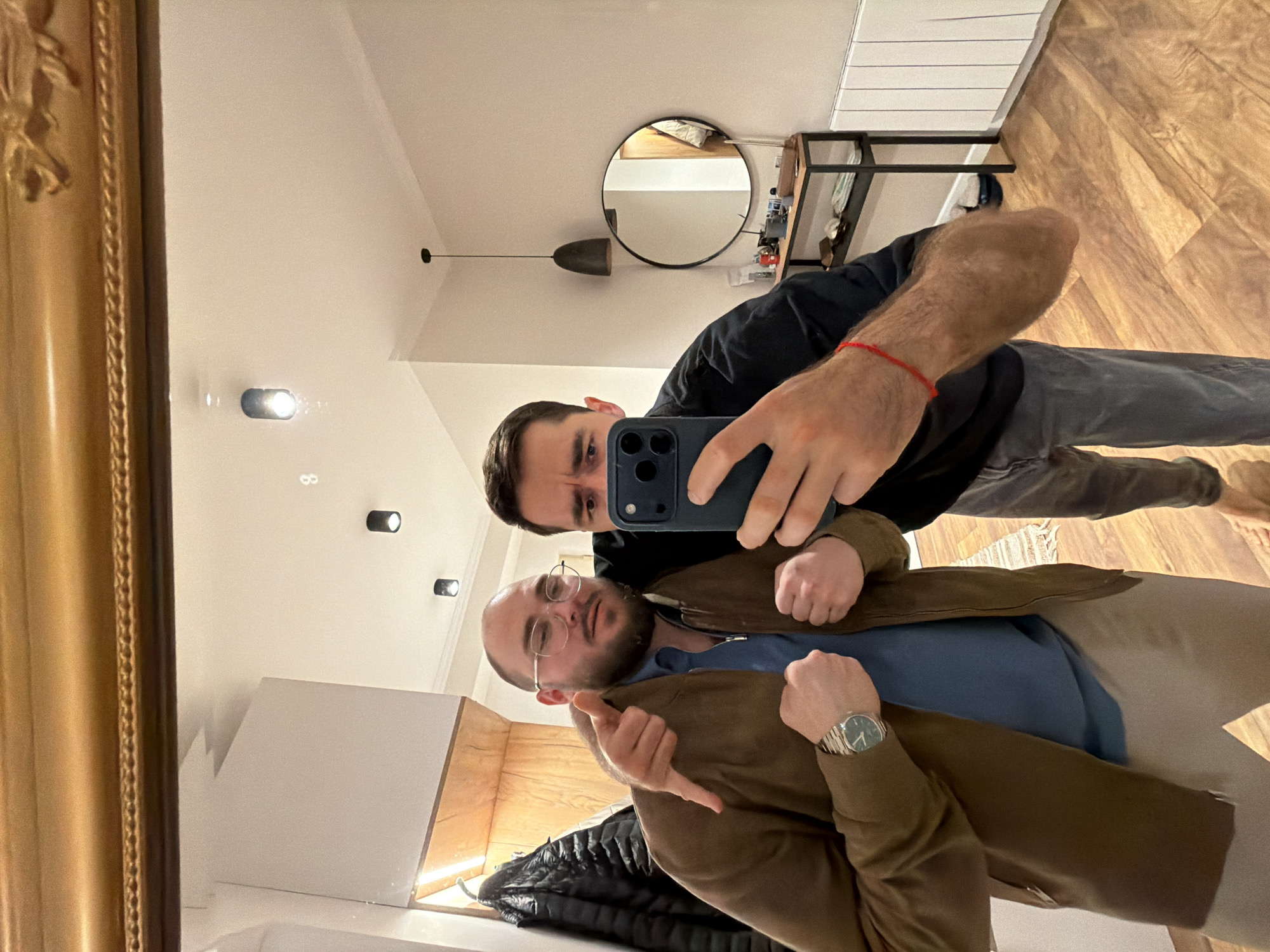 Two men are taking a selfie in a mirror. The man holding the phone is wearing a black shirt and grey jeans. The other man is wearing glasses, a blue shirt, a brown jacket, and a watch. They appear to be indoors, possibly in a hallway or entryway. There is a mirror on the wall with a black frame, a console table, and a wooden floor. The location is likely Tbilisi, Georgia.